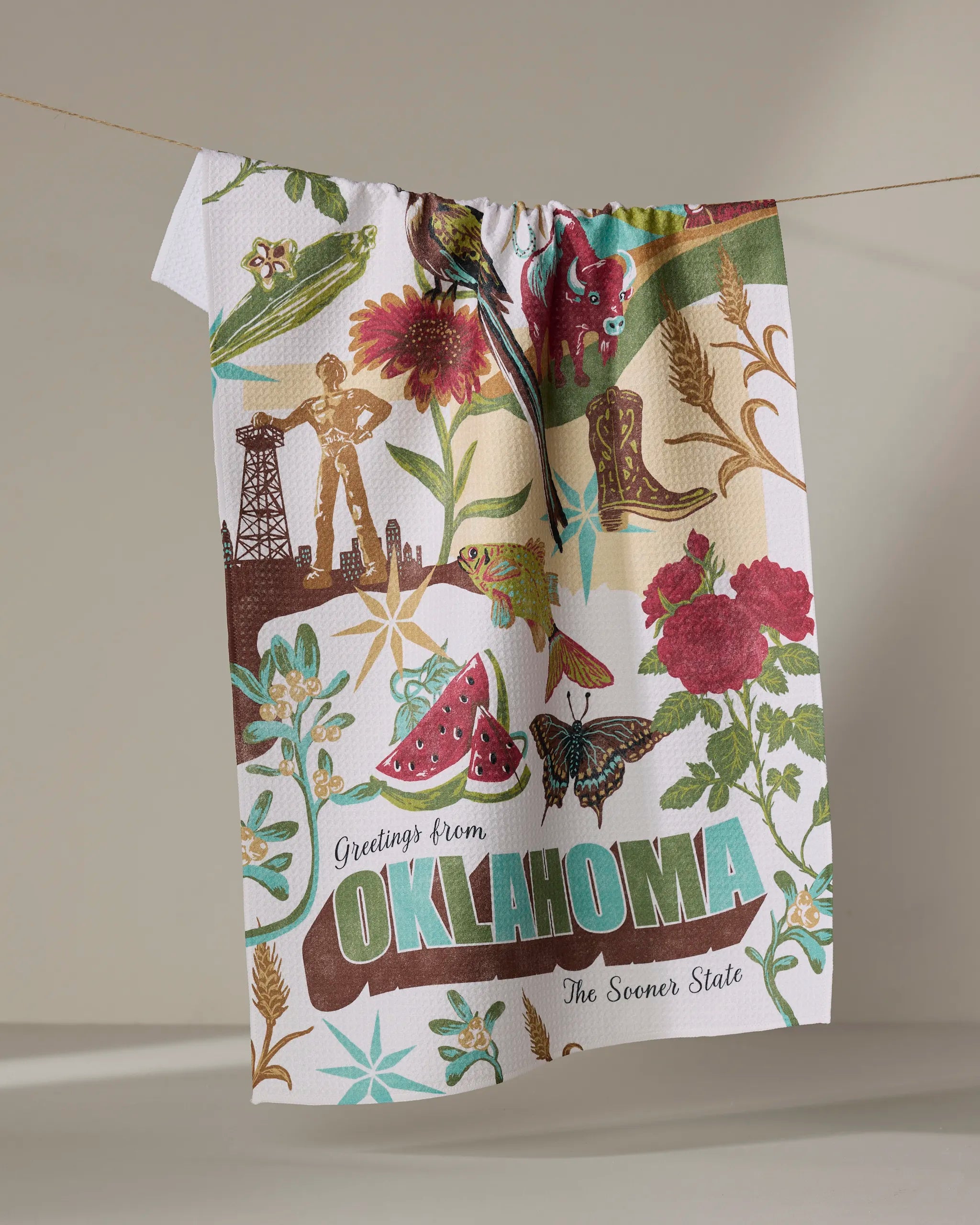 The Geometry "Greetings From Oklahoma" tea towel hangs to dry, vibrant with quick-drying fabric and illustrations of state icons—an oil rig, cowboy boots, flowers, butterfly, watermelon—and the phrase “Greetings from Oklahoma, The Sooner State.”.
