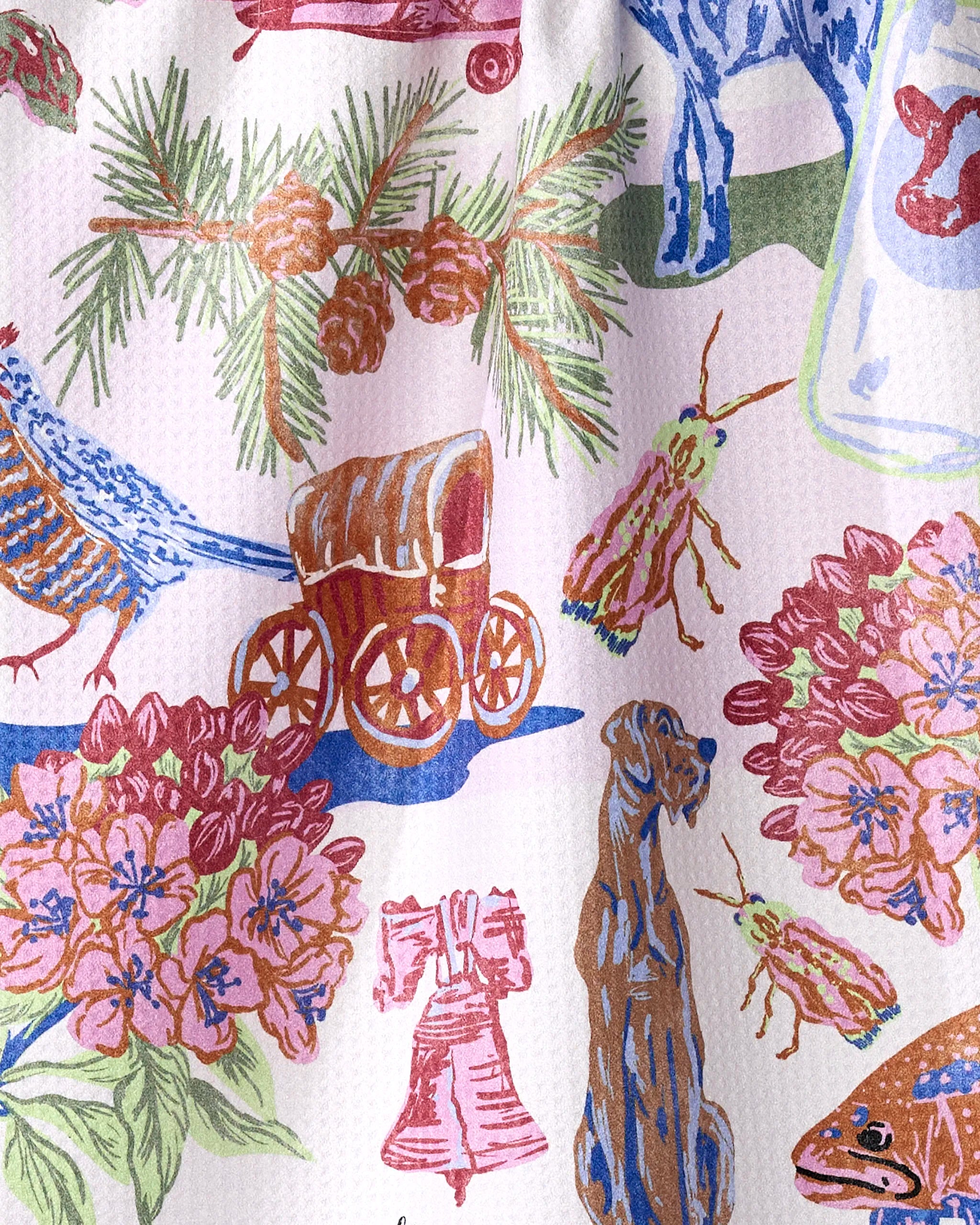 The Geometry "Greetings From Pennsylvania" tea towel features a colorful pattern with a covered wagon, pine branches, cones, and a blue dog on a white textured background. Made from recycled materials, it’s quick-drying and vibrant.