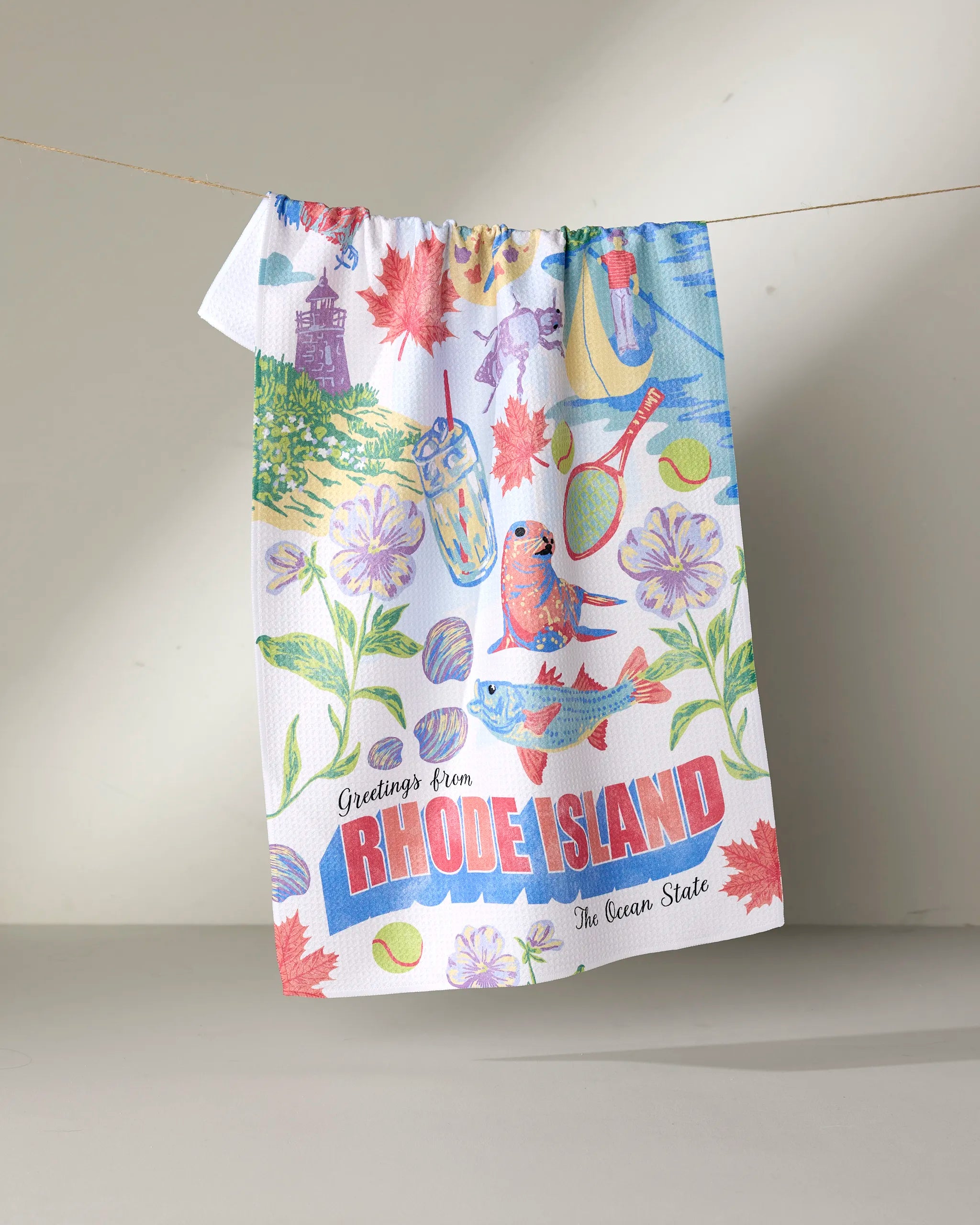 The Geometry "Greetings From Rhode Island" tea towel, made from recycled materials, features vibrant flower, bird, drink, and Rhode Island icon illustrations along with the text "Greetings from RHODE ISLAND, The Ocean State.