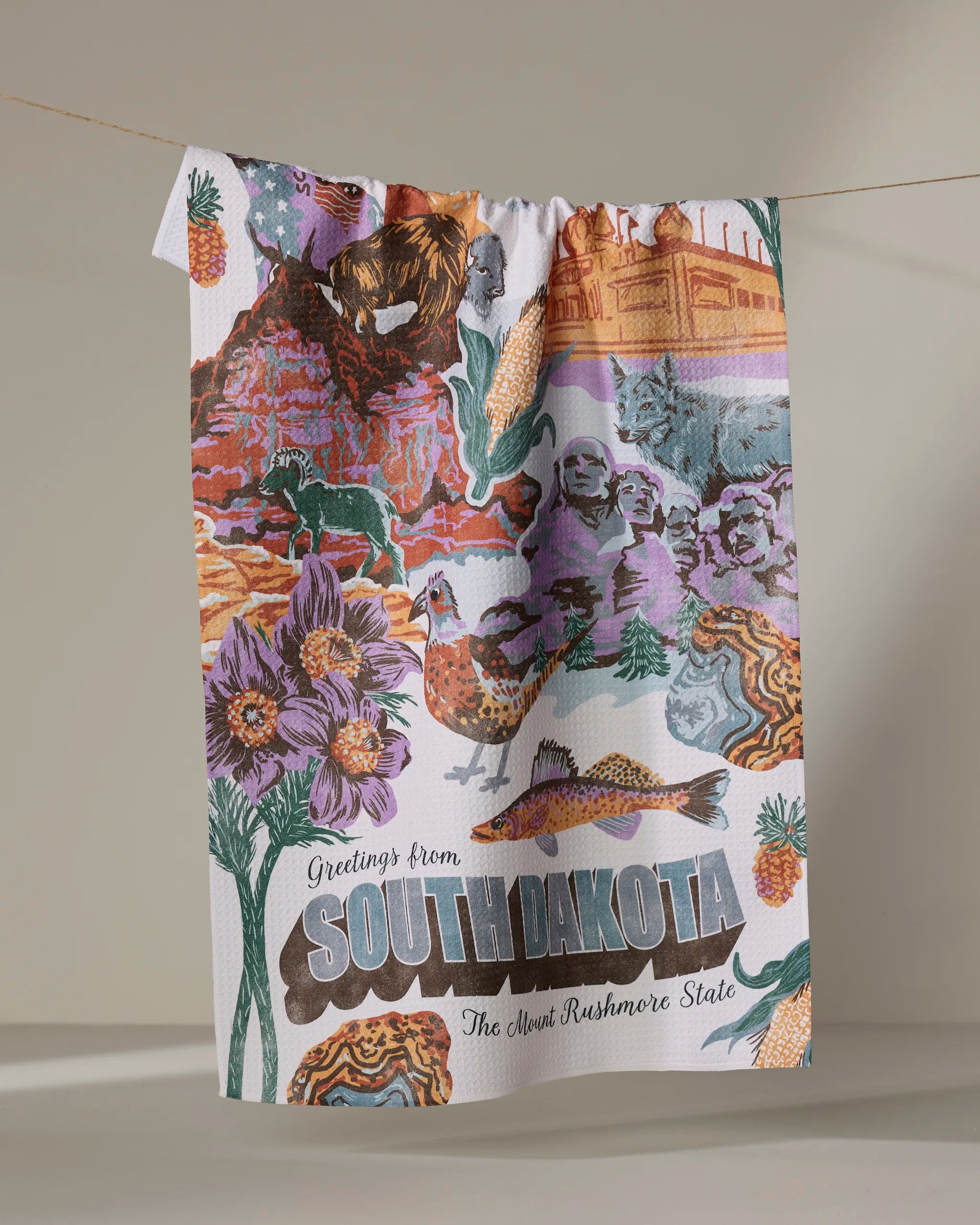 The Geometry "Greetings From South Dakota" tea towel, made from quick-drying recycled materials, showcases vibrant illustrations of Mount Rushmore, bison, wildflowers, and “Greetings from South Dakota, The Mount Rushmore State.”.