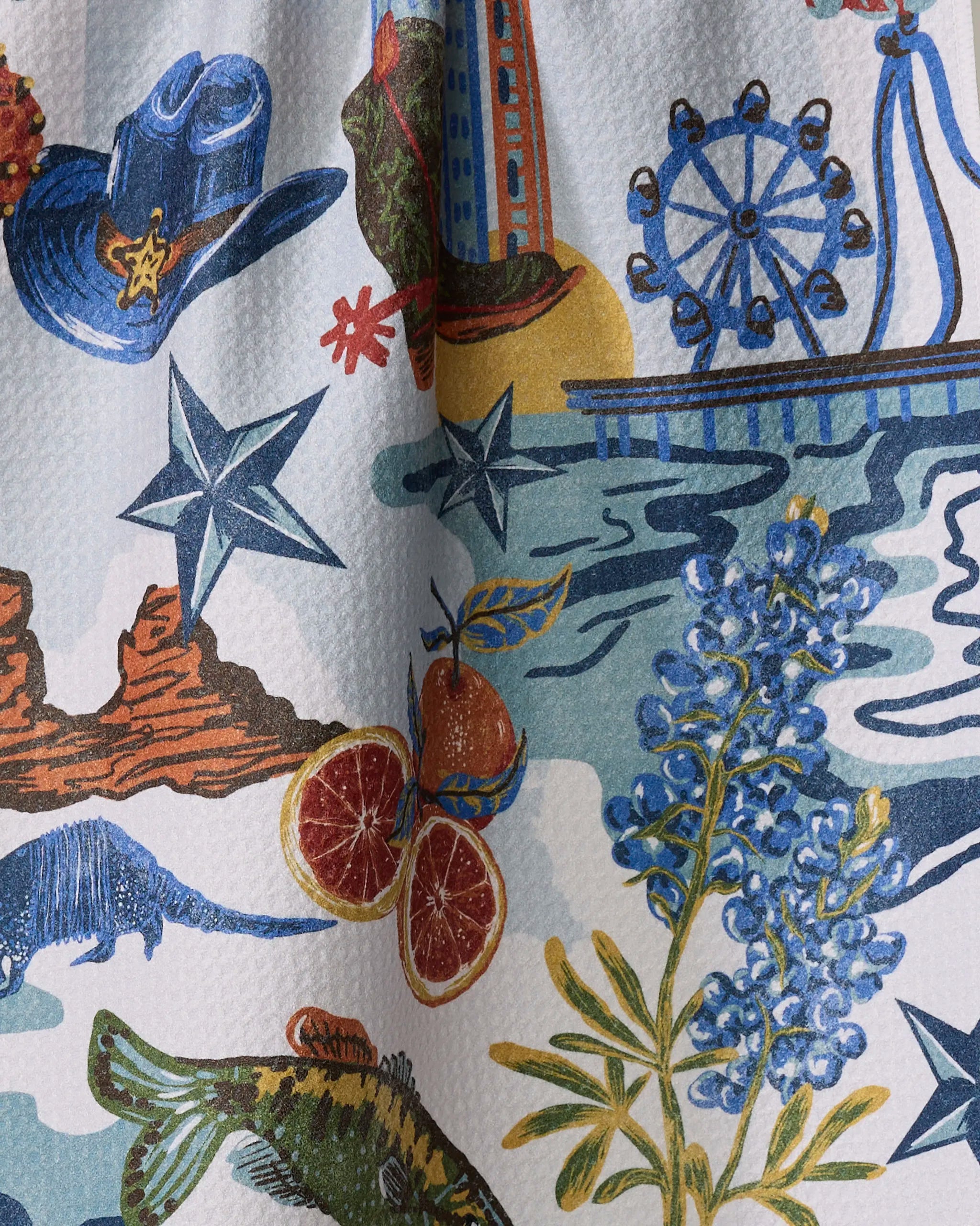 Geometry’s “Greetings From Texas” tea towel showcases a vibrant pattern of Texas icons—cowboy hat, ferris wheel, bluebonnets, citrus, fish, stars, desert rocks—printed on quick-drying textured fabric.