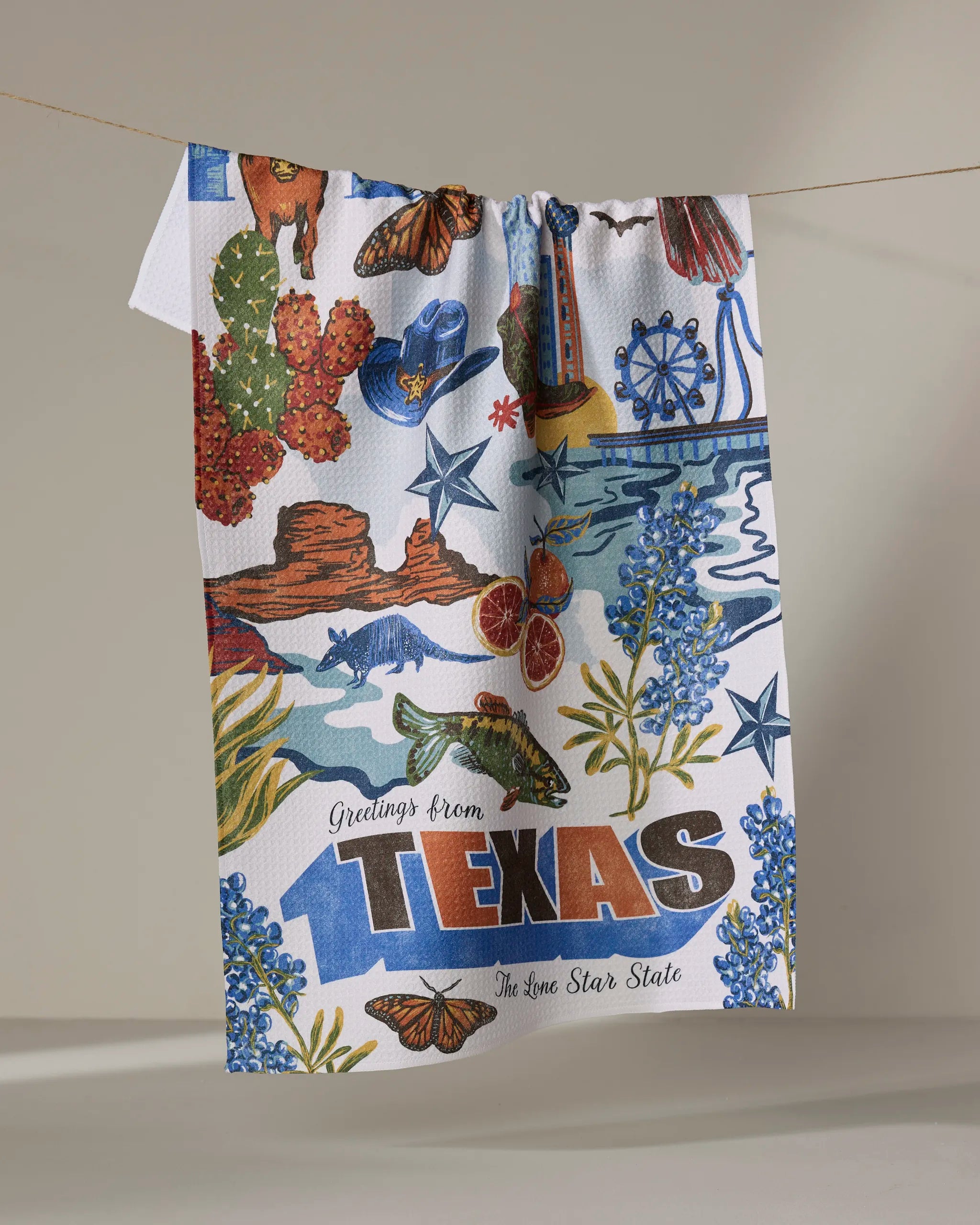The Geometry "Greetings From Texas" tea towel showcases cacti, longhorns, bluebonnets, an armadillo, and a Ferris wheel, plus “Greetings from Texas, The Lone Star State.” Made from quick-drying, recycled materials.