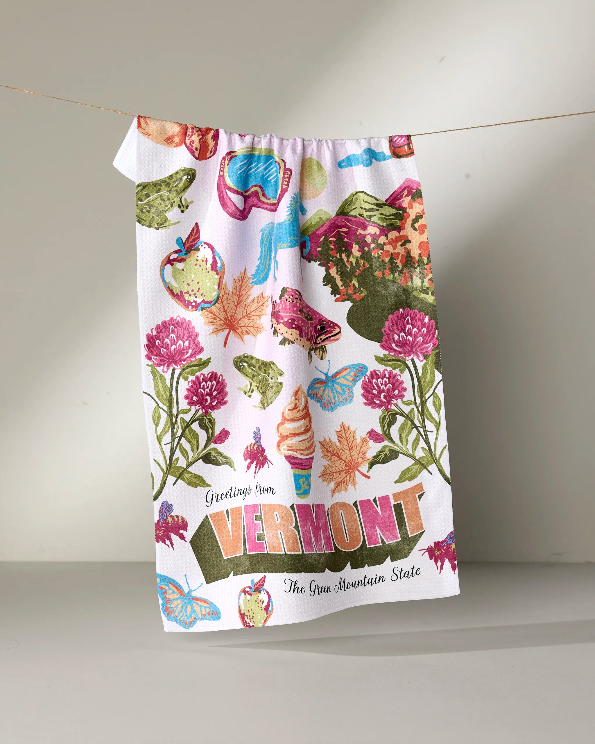 The Geometry "Greetings From Vermont" kitchen towel, made from recycled materials, features colorful illustrations of Vermont icons—flowers, maple leaves, a covered bridge, ice cream, and mountains.