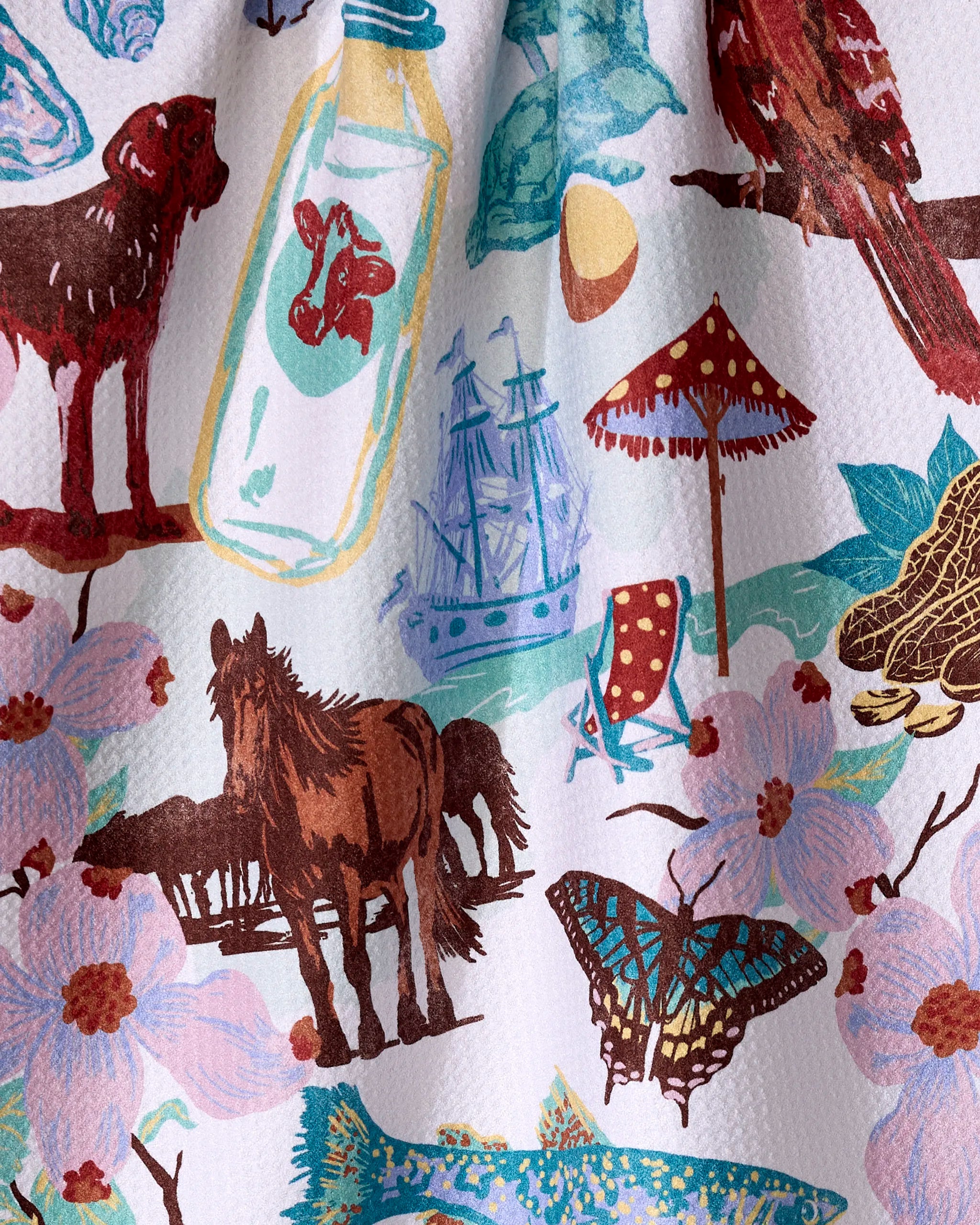 The "Greetings From Virginia" tea towel by Geometry features a vibrant illustration of local icons—a horse, butterfly, dog, lobster bottle, pirate ship, umbrella with chair, tree, birds, flowers, turtle, and lemon—on quick-dry recycled fabric.