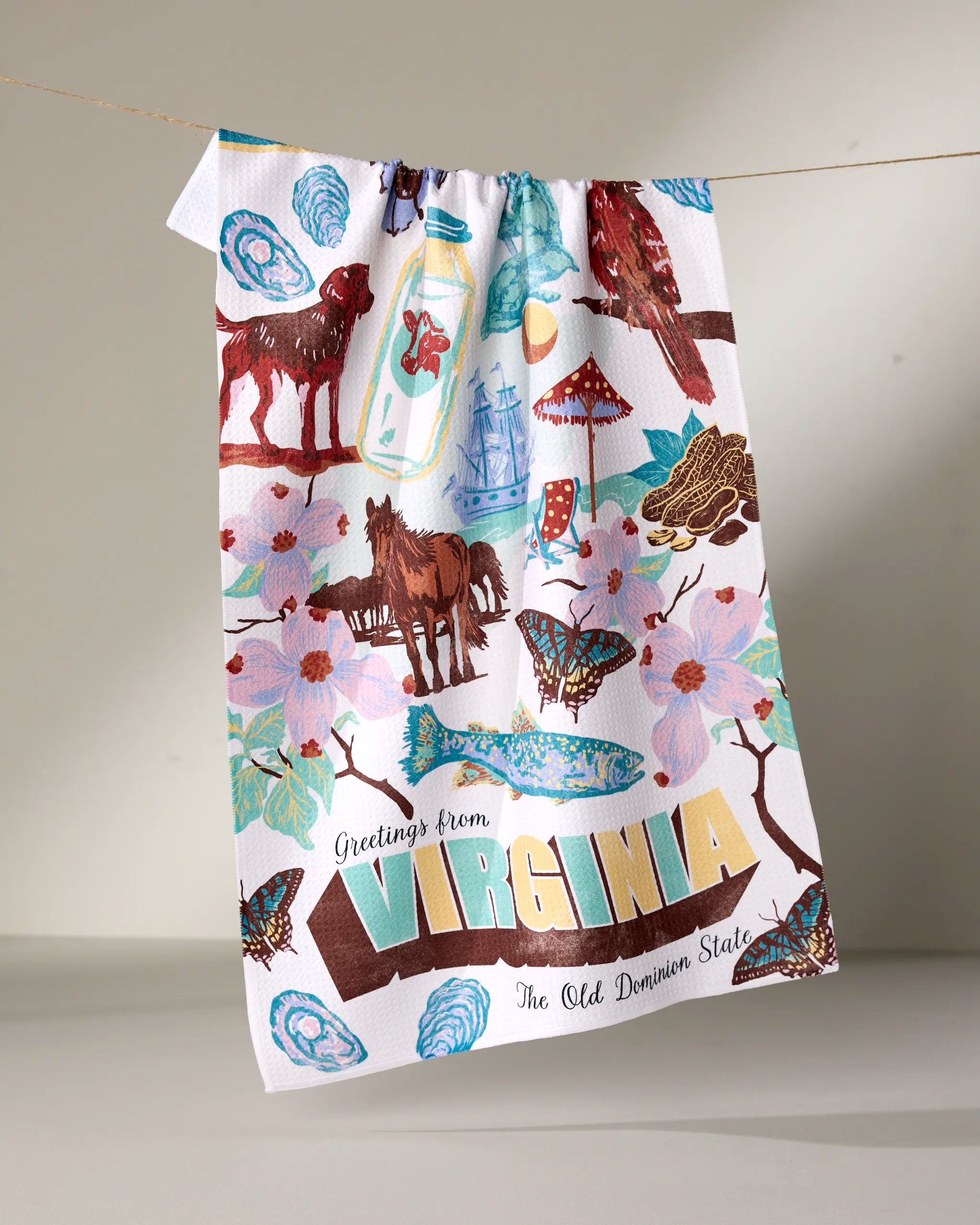 The Geometry "Greetings From Virginia" tea towel showcases vibrant illustrations and text, featuring animals, flowers, butterflies, and Virginia icons. Made from quick-drying recycled materials for both style and sustainability.
