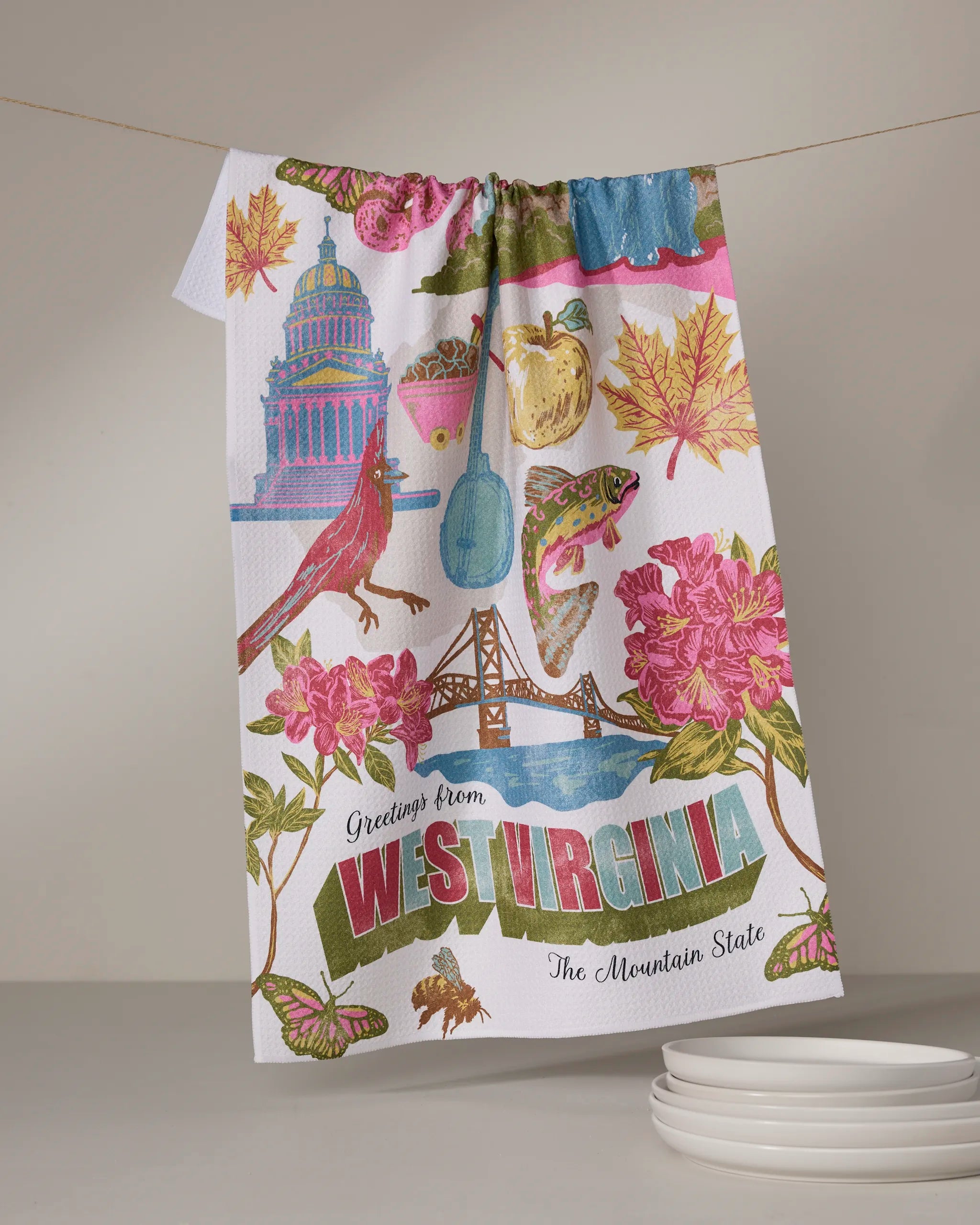 The Geometry "Greetings From West Virginia" tea towel features colorful state symbols—Capitol, cardinal, trout, rhododendron, and a bridge. Made from recycled materials, it reads "Greetings from West Virginia" at the bottom.