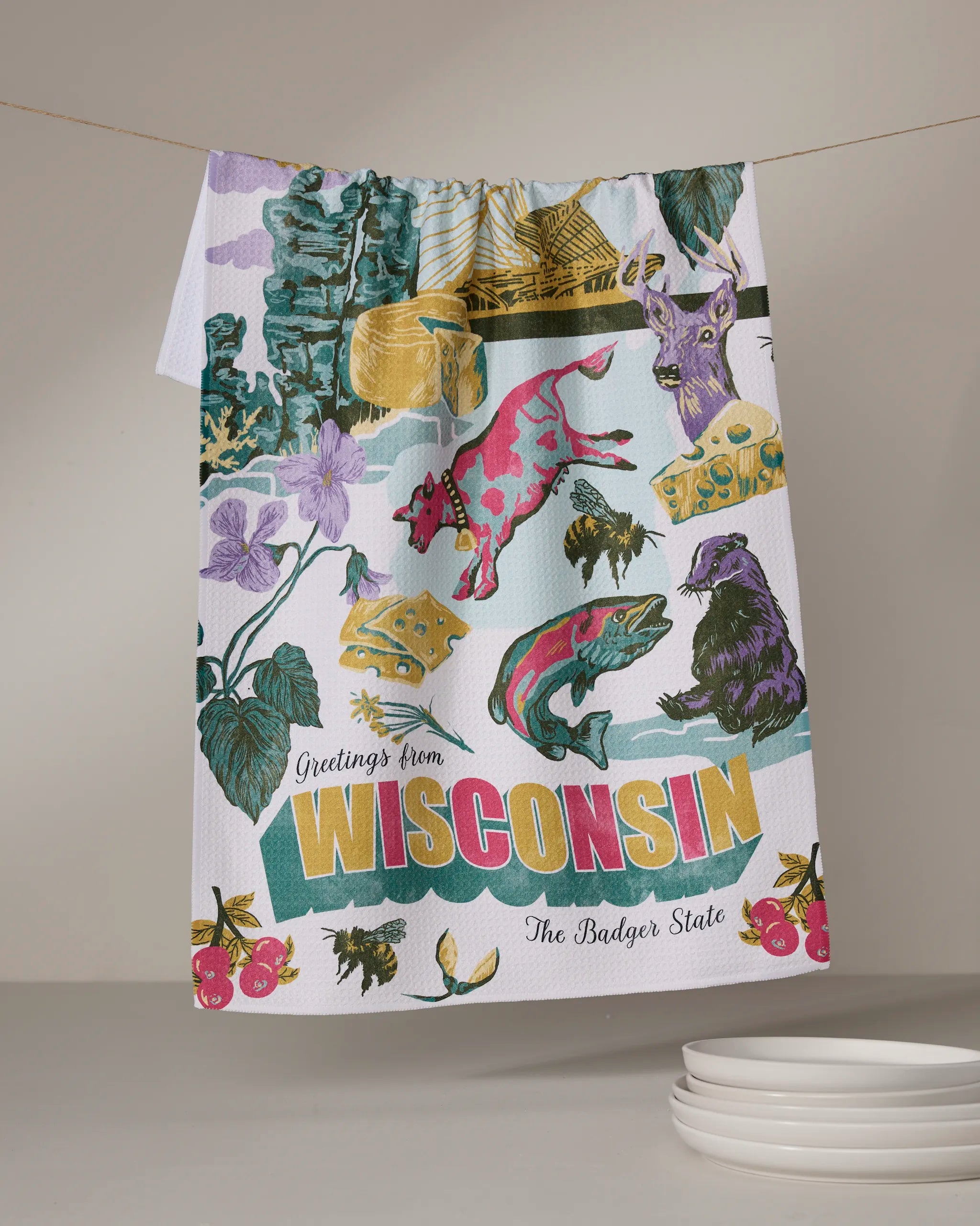 The Geometry "Greetings From Wisconsin" tea towel, made from recycled materials, features colorful Wisconsin symbols—cow, cheese, deer, flowers, and badger—with "Greetings from Wisconsin The Badger State" text. Plates are stacked below.