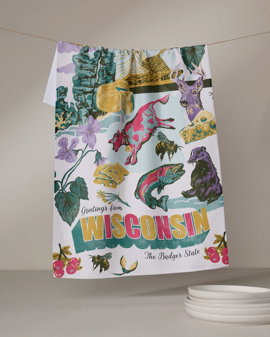 The Geometry "Greetings From Wisconsin" tea towel, made from recycled materials, features colorful Wisconsin symbols—cow, cheese, deer, flowers, and badger—with "Greetings from Wisconsin The Badger State" text. Plates are stacked below.