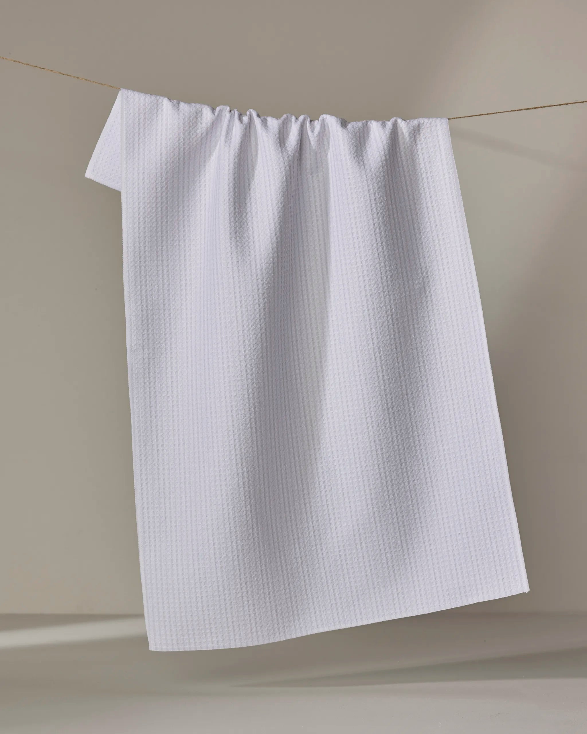 The Grey Salt towel by Kailee Wright, featuring a textured quick-dry design, hangs neatly on a clothesline against a softly lit beige background.