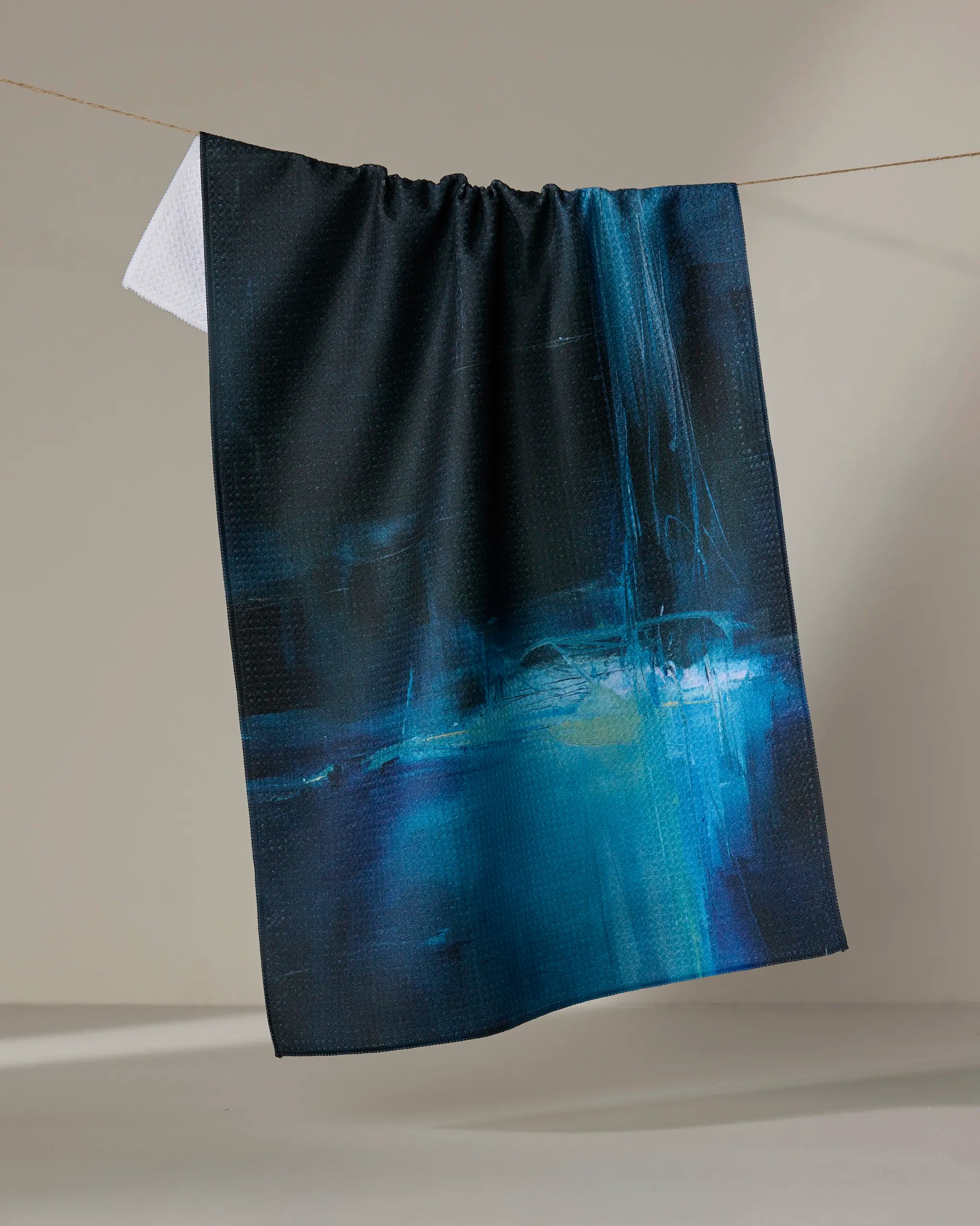 The Grotto kitchen tea towel by Jennifer Janesko, featuring an abstract blue and black design, hangs on a string against a neutral background with the white reverse side visible.