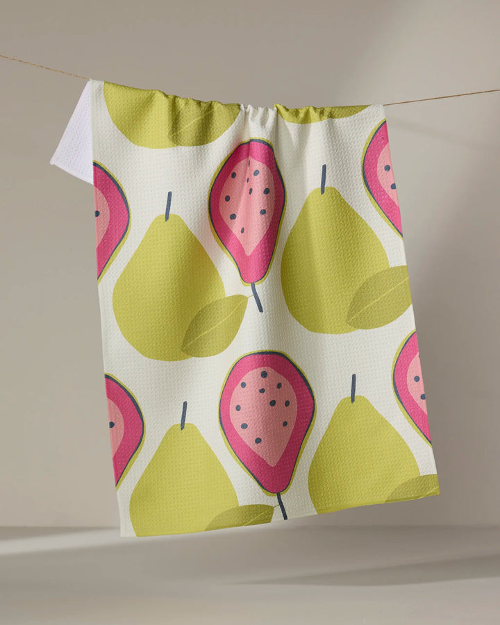 The Lizzie Clark "Guava Groove" recycled kitchen towel, adorned with yellow pears and pink guavas, hangs on a string against a neutral background.