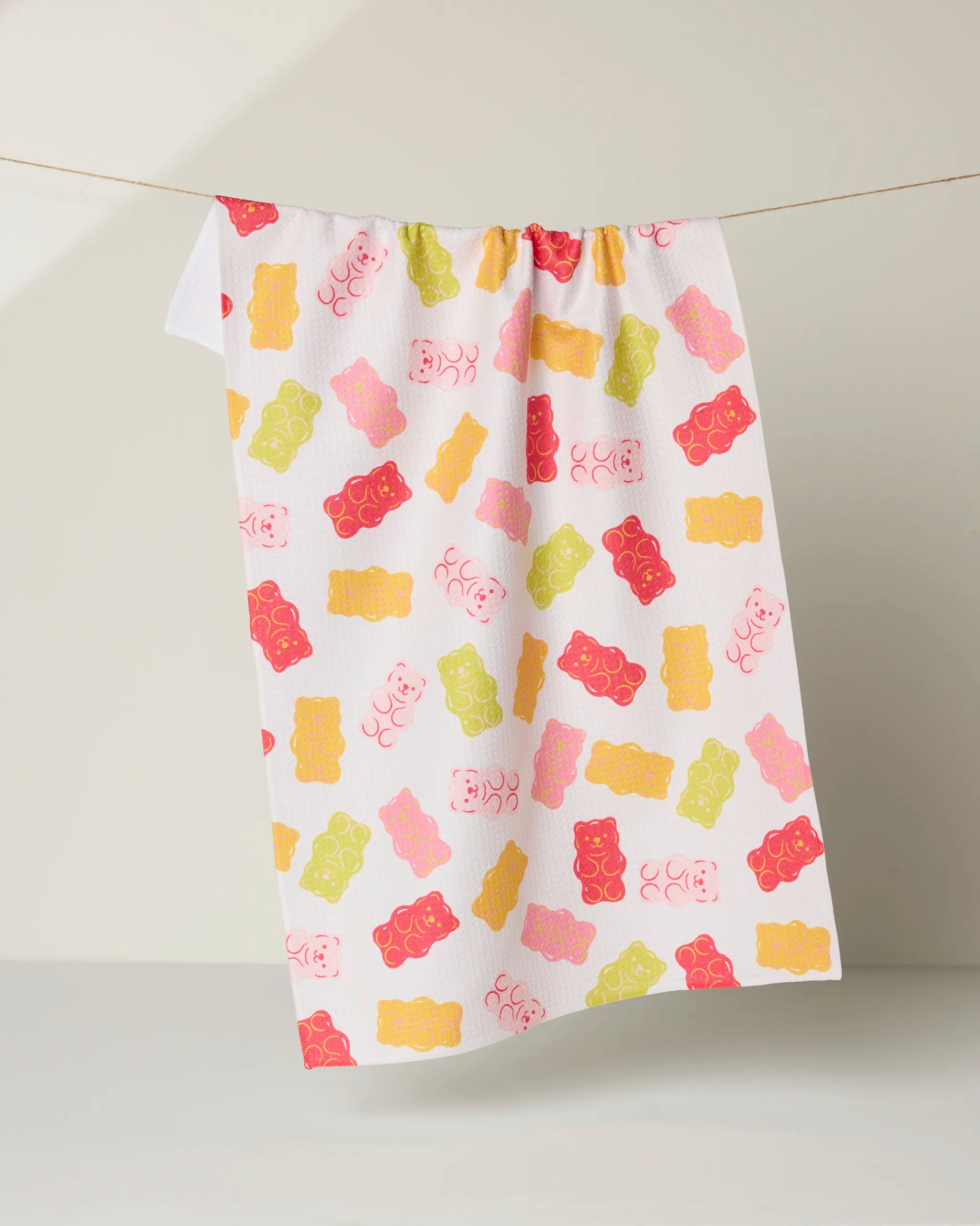 The Gummy Bear tea towel by Vaishali Design Studio is ultra-absorbent, white, and features a playful pattern of pink, yellow, green, and red gummy bears hanging against a light background.