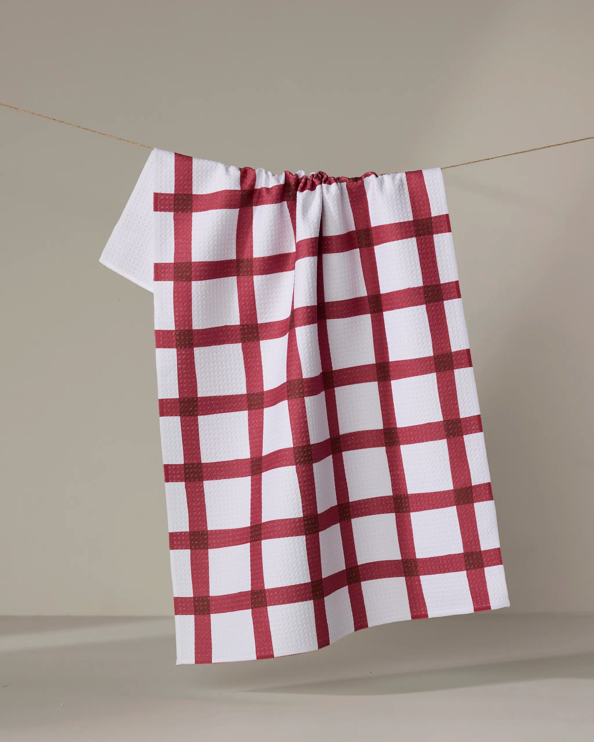 The Amy MacCready Hackett is a white tea towel with a red checkered pattern, made from recycled materials, shown hanging on a clothesline against a plain, light background.