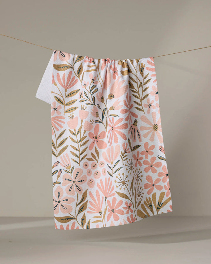 The "Halia" recycled kitchen towel by Amy MacCready, featuring pink and gold floral and leaf patterns, hangs on a clothesline against a plain beige background.
