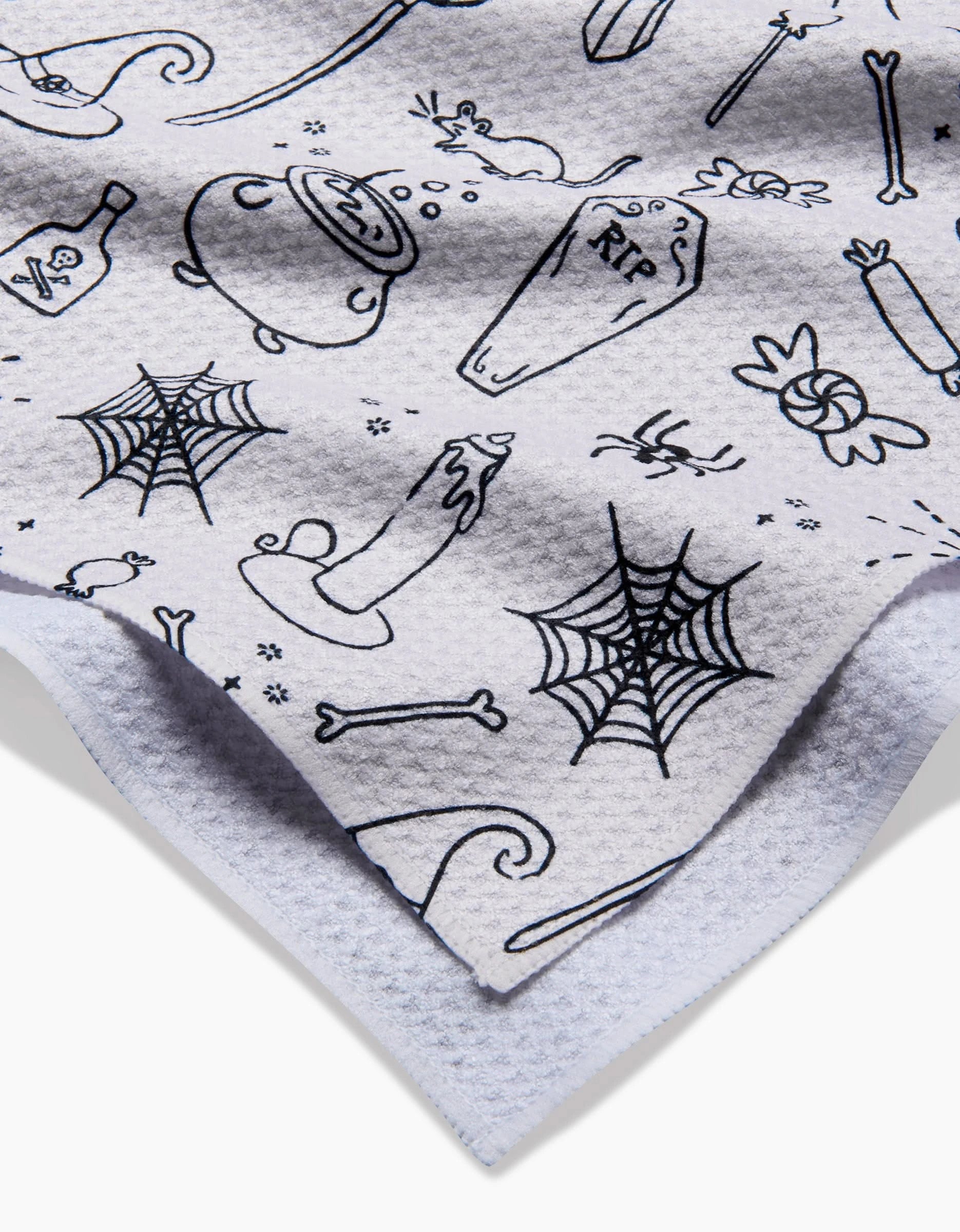 A close-up of Kayla Anns Halloween Magic White shows ultra-absorbent Geoweave fabric with black Halloween-themed drawings—spiderwebs, candles, cauldrons, candy, bones, a RIP coffin, rats, and magic wands—highlighting its double-layer design.