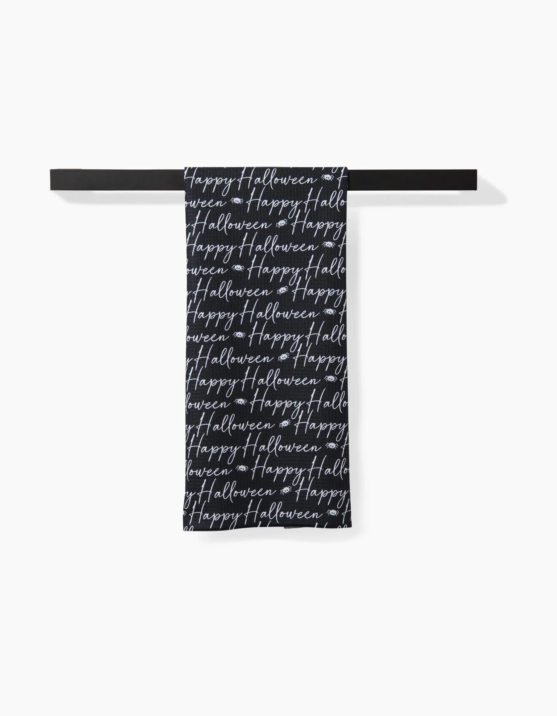 The Geometry Halloween Script Black towel, featuring Happy Halloween in white cursive, is ultra-absorbent and hangs folded over a black bar against a white background.