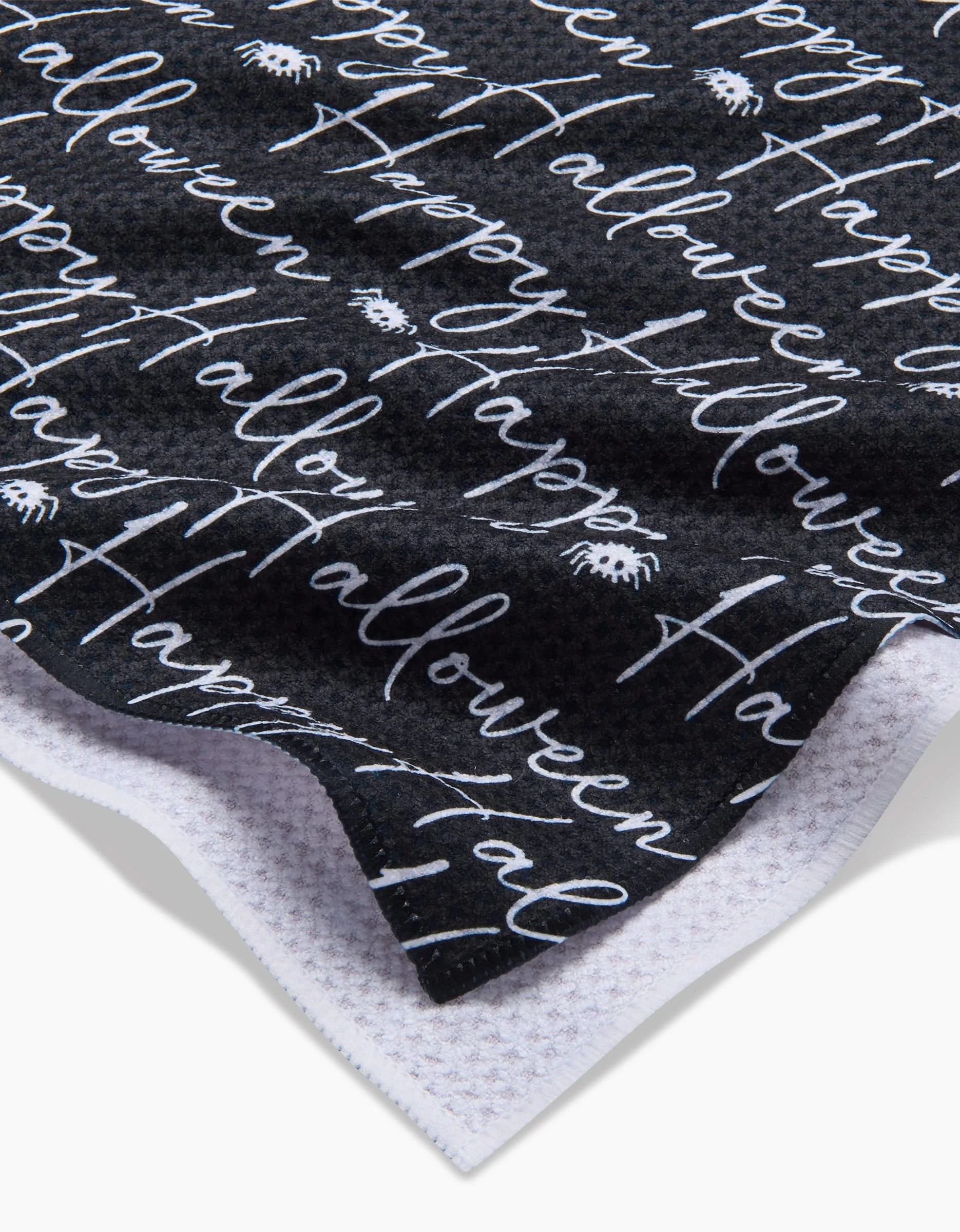 A close-up of the Geometry Halloween Script Black recycled kitchen towel featuring white Happy Halloween script and spider illustrations, highlighting its textured fabric and white underside.