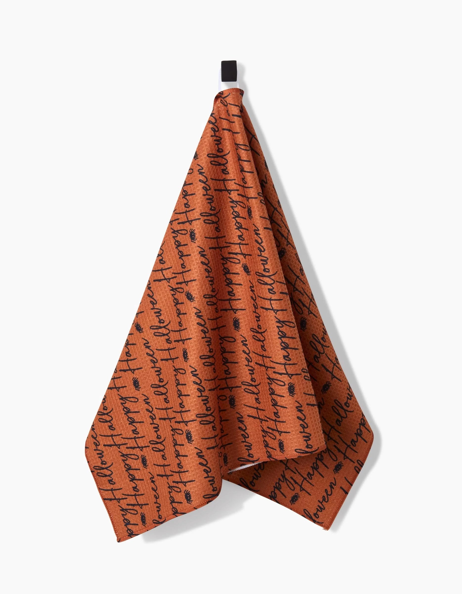 The Geometry Halloween Script Orange tea towel, made from recycled materials, displays black handwritten script and dot patterns on rust-orange fabric and hangs from a small black loop against a white background.