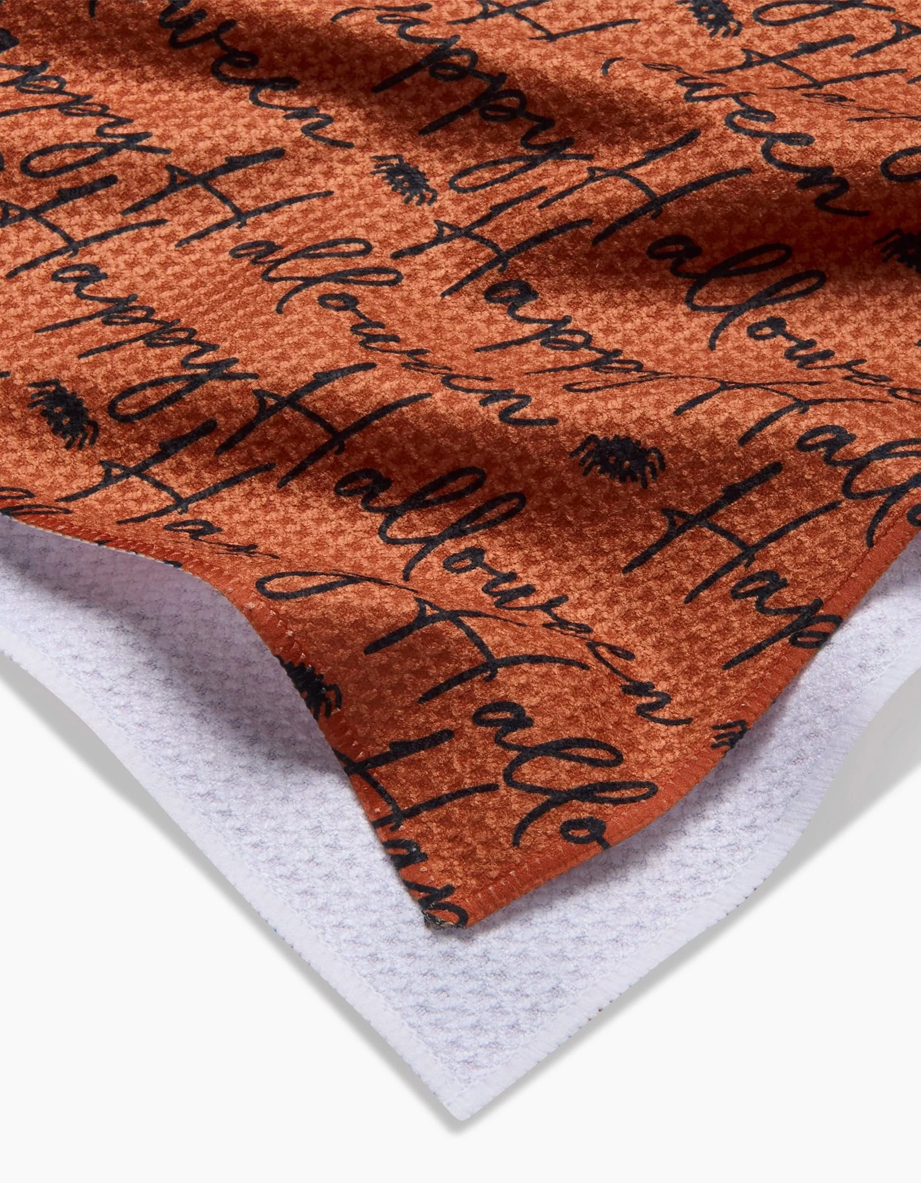Close-up of the Geometry Halloween Script Orange kitchen towel, made from recycled materials, featuring Happy Halloween in black script with small black spider illustrations, layered over white textured fabric.