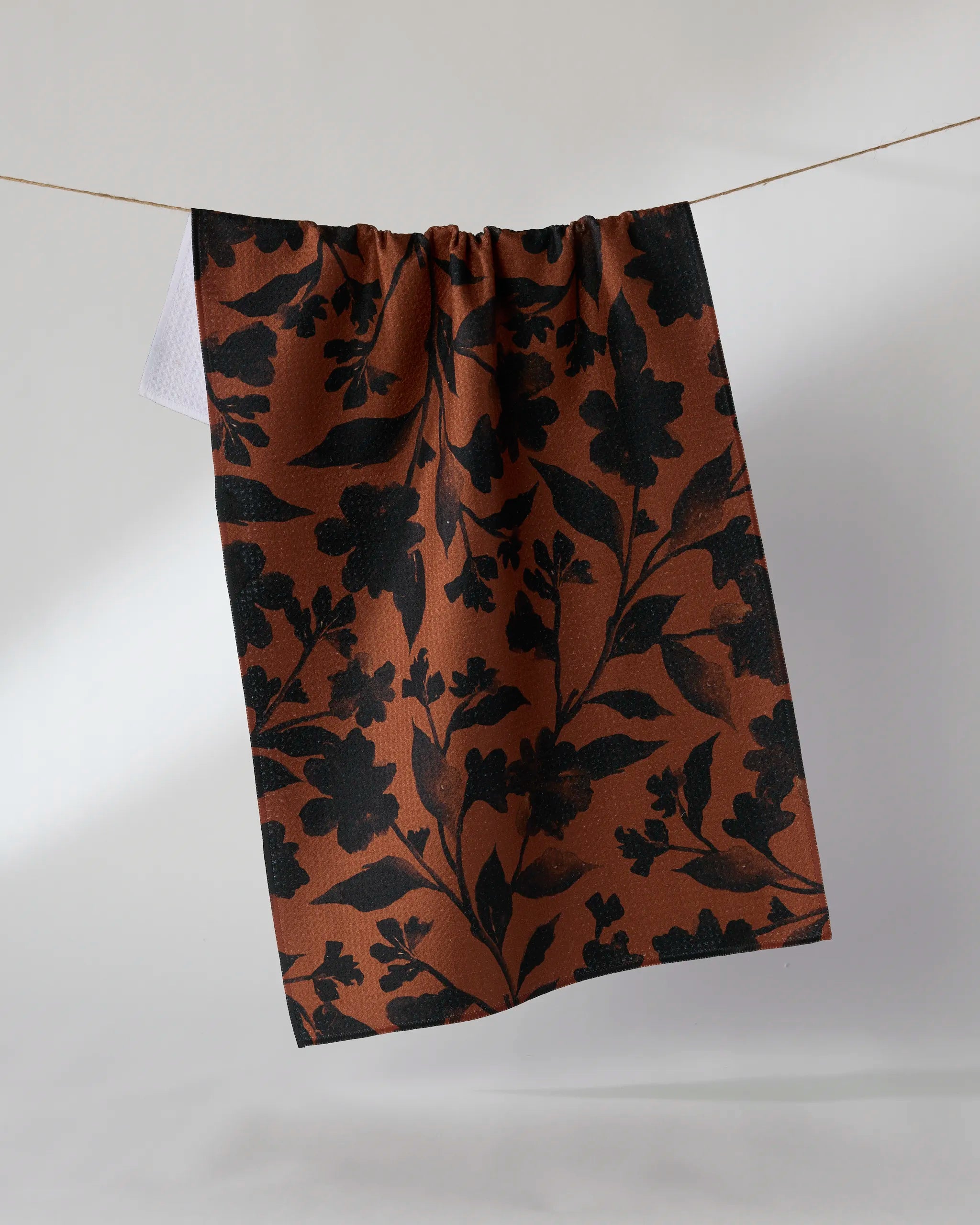 The Geometry Hallowell is a brown kitchen tea towel with a black floral pattern, made from recycled materials and shown hanging on a clothesline against a light background.