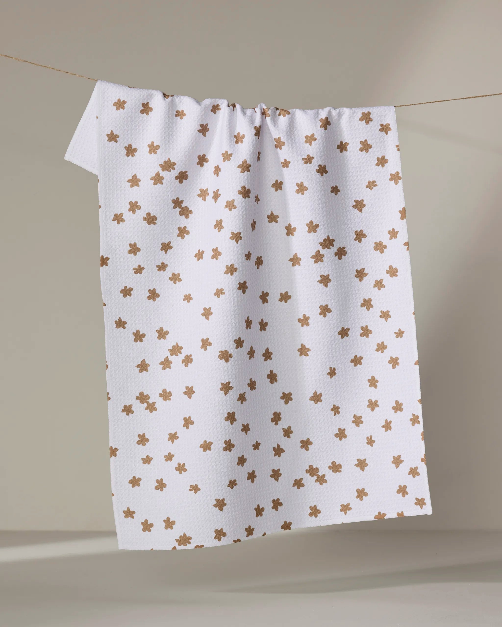 The Julianne Haness "Haness Golden" is a white, absorbent dish towel with a textured pattern and light brown floral prints, shown hanging on a thin string against a neutral background.