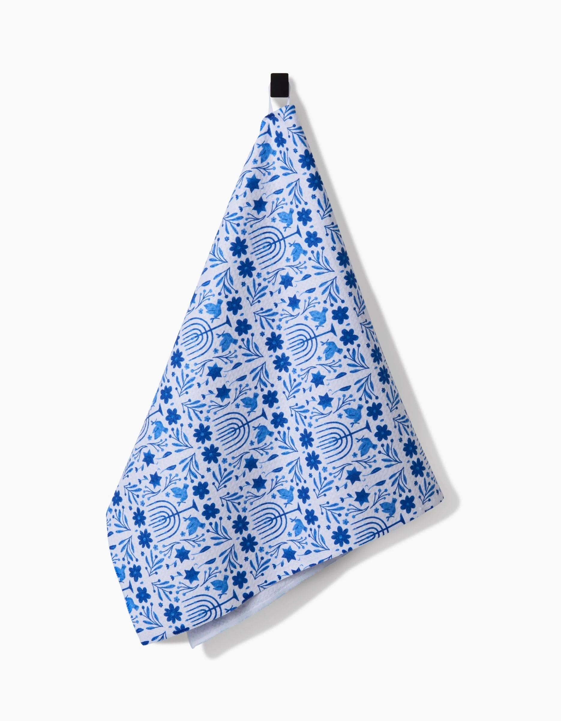 The Laura Supnik Hanukkah kitchen tea towel, featuring blue and white floral and geometric patterns and crafted from recycled materials, hangs by a black loop against a plain white background.