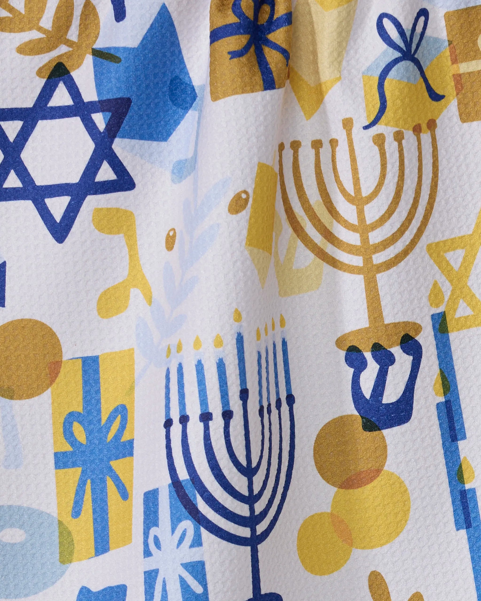 The "Hanukkah is Here" kitchen towel by Geometry features blue and gold menorahs, Stars of David, dreidels, gelt, gifts, and olive branches on a textured white background—eco-friendly and made from recycled materials.