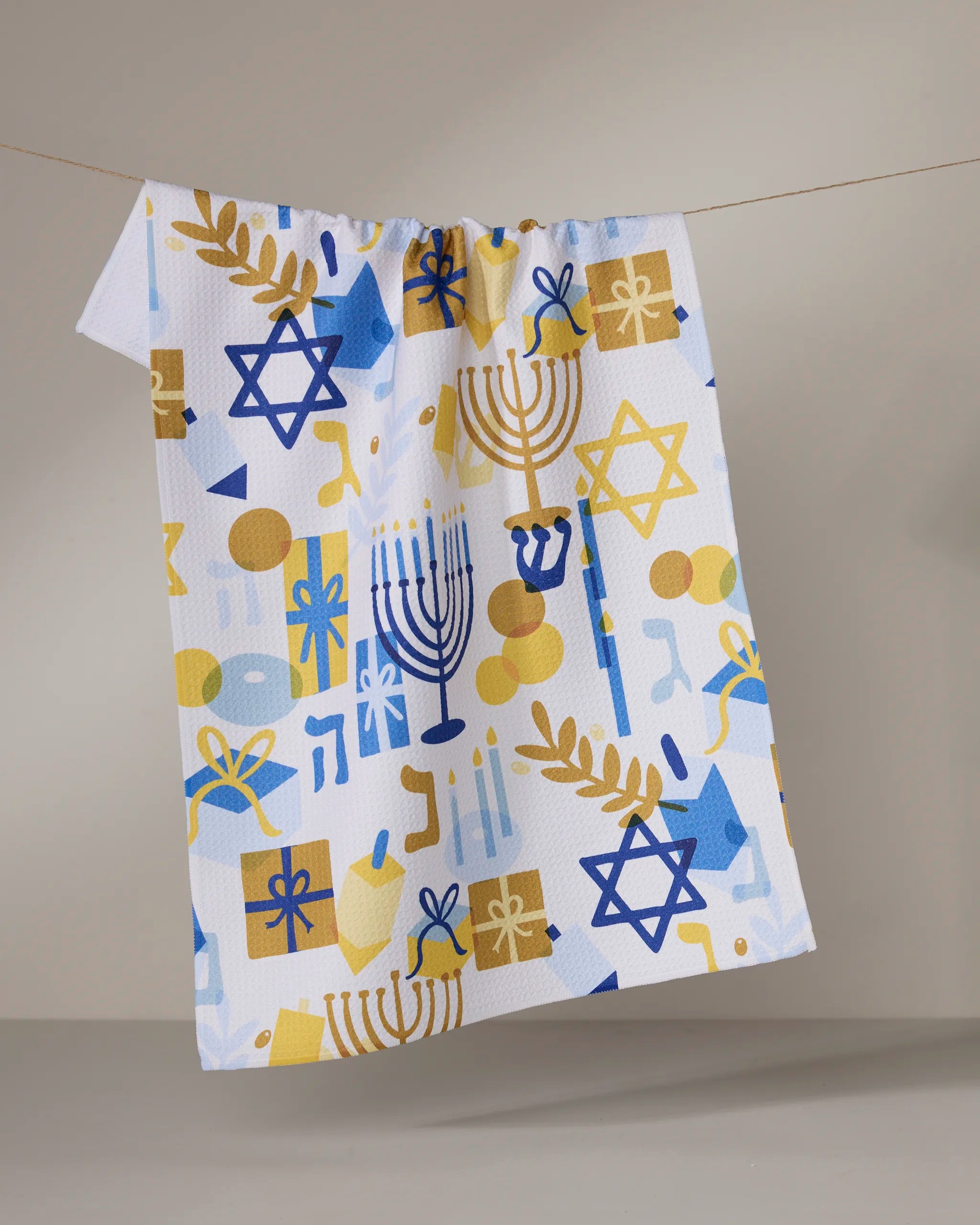The "Hanukkah is Here" tea towel by Geometry features blue and gold Stars of David, menorahs, dreidels, and gelt on recycled fabric, shown hanging on a clothesline against a plain background.