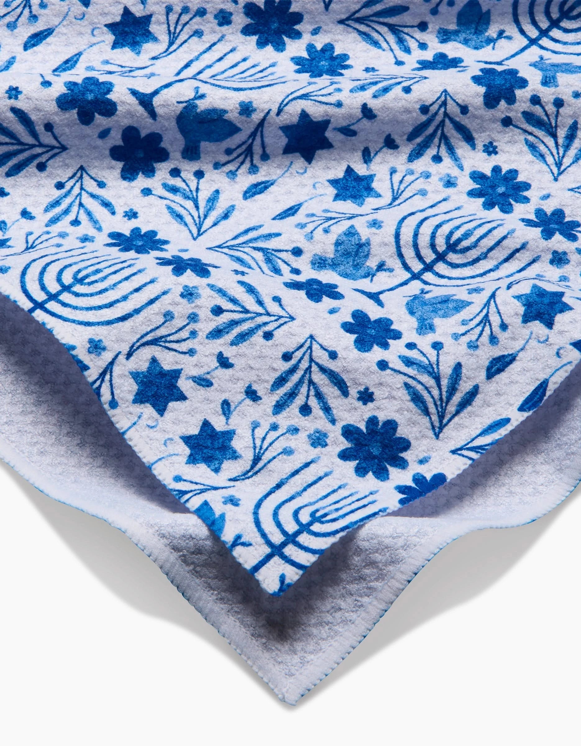 A close-up of Laura Supniks Hanukkah tea towel, crafted from recycled materials. The soft white fabric features blue menorahs, Stars of David, dreidels, and flowers, with slightly wavy edges for added texture.
