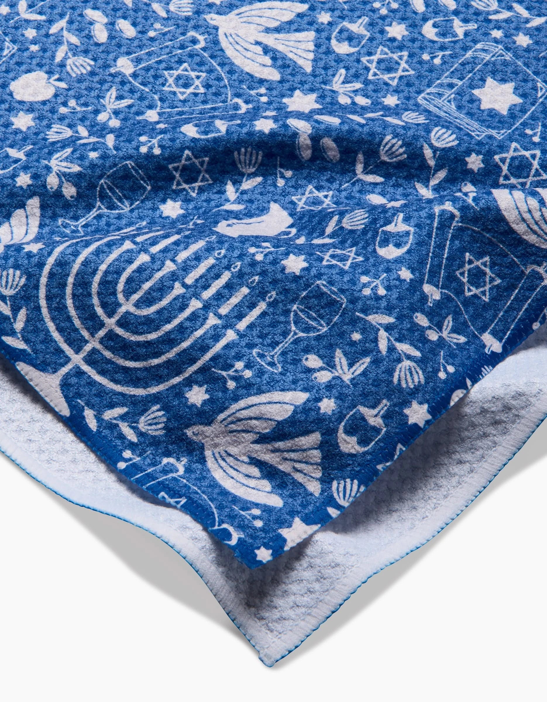 The Hanukkah Sameach by Megan Kampa is a blue and white blanket featuring menorahs, dreidels, Stars of David, and more—perfect as a cozy throw or quick-drying kitchen towel, with both colorful sides beautifully displayed.