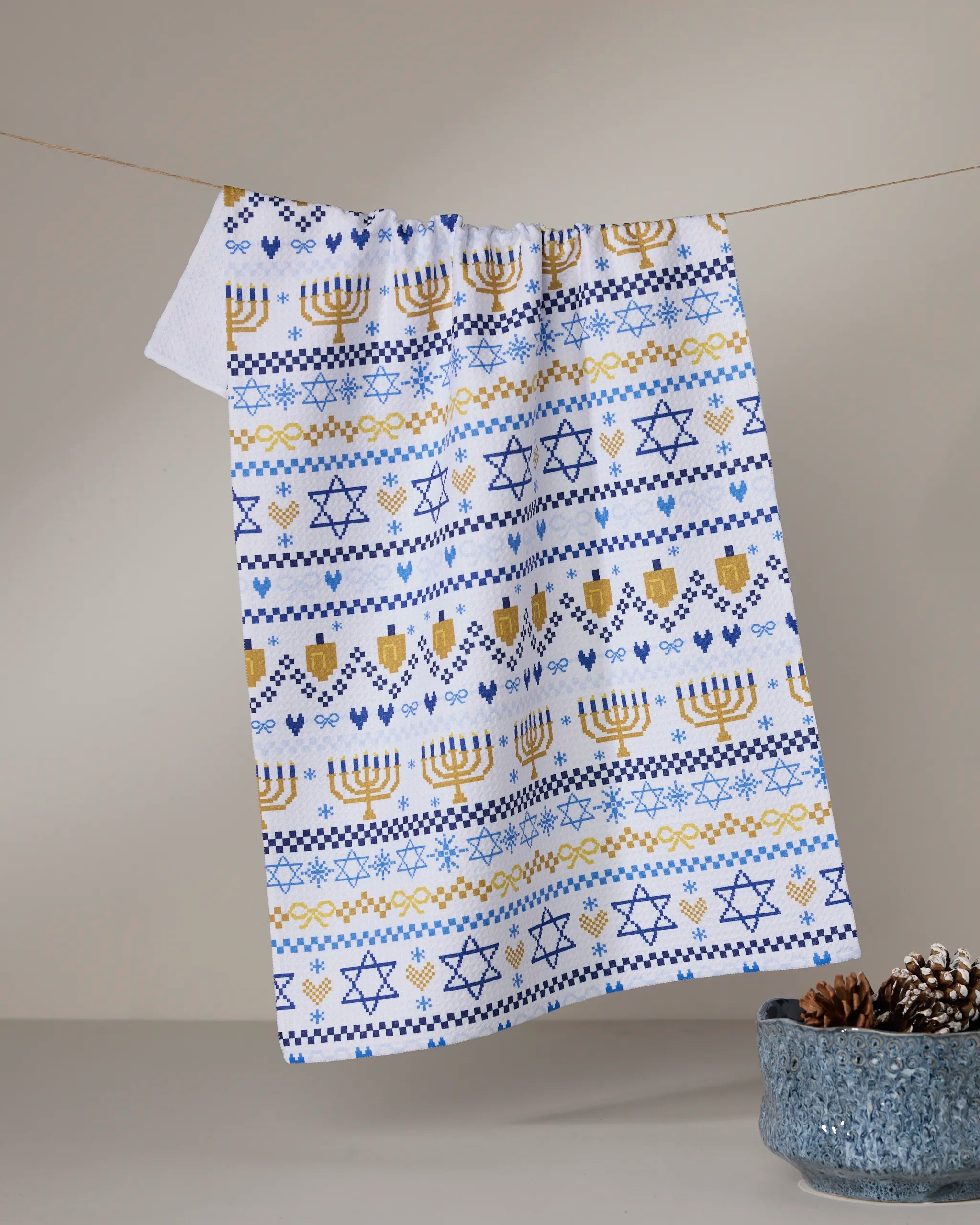 The Geometry Hanukkah Sweater towel, featuring blue and gold menorahs, dreidels, Stars of David, and hearts on white eco-friendly fabric, hangs on a string above a metal bucket filled with pinecones.