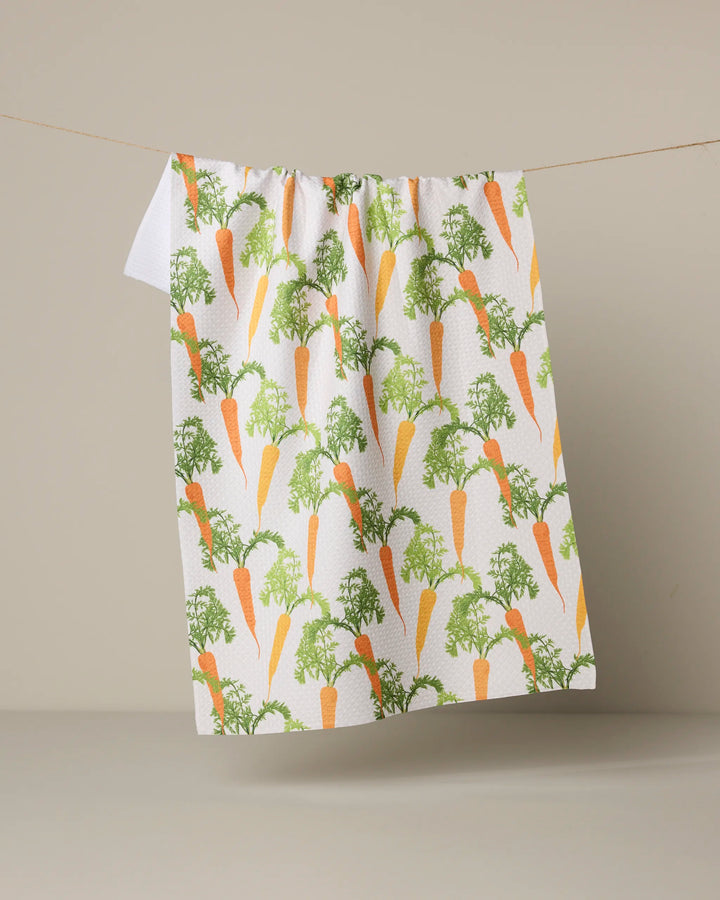 The Geometry "Happy Carrots" tea towel, featuring a white background with repeating orange carrot and green leaf patterns, hangs on a string against a plain beige backdrop.