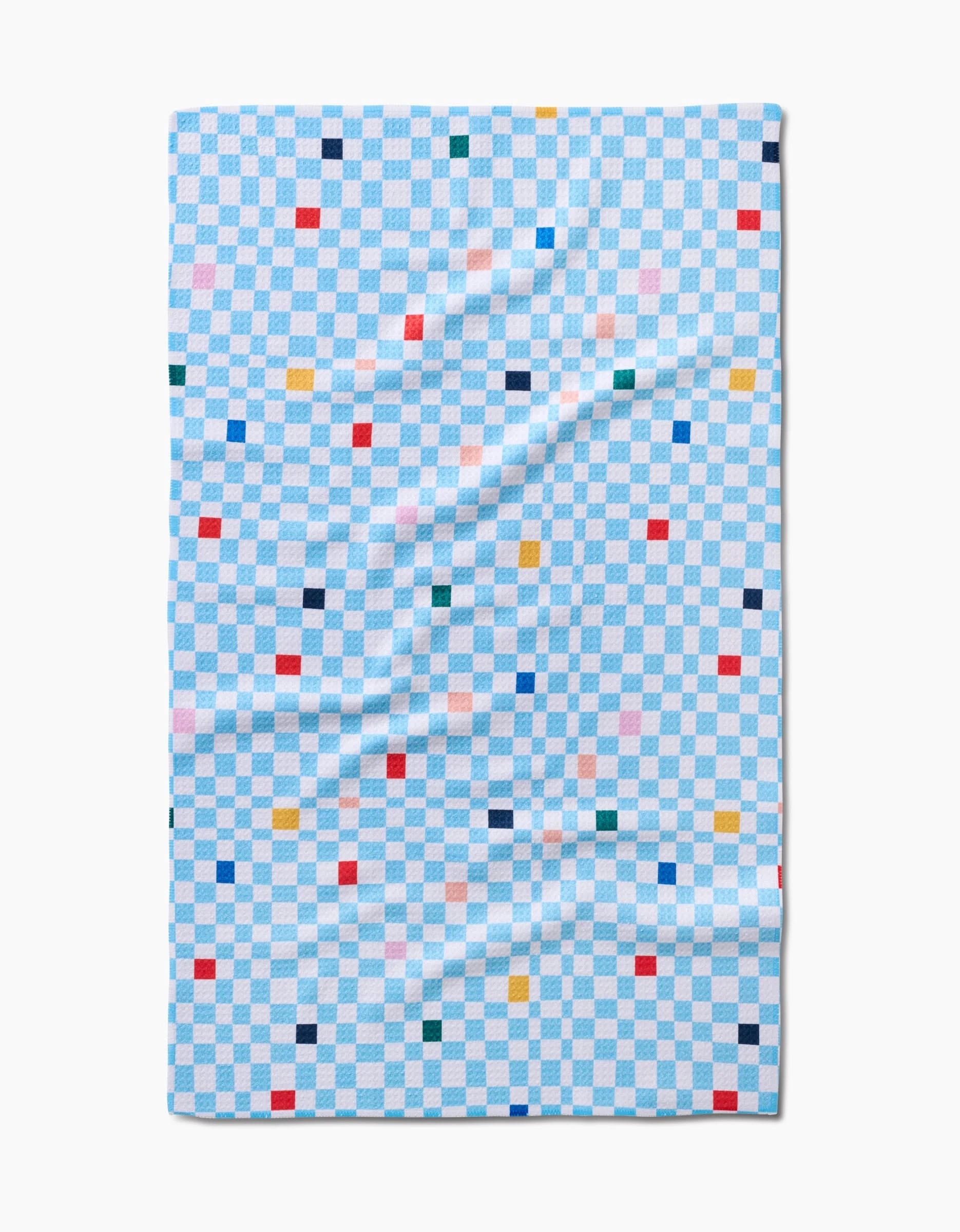 The Happy Hemera Happy Check tea towel, made from ultra-absorbent Geoweave™ fabric, features a light blue and white checkerboard with colorful squares. It’s slightly wrinkled and laid flat.
