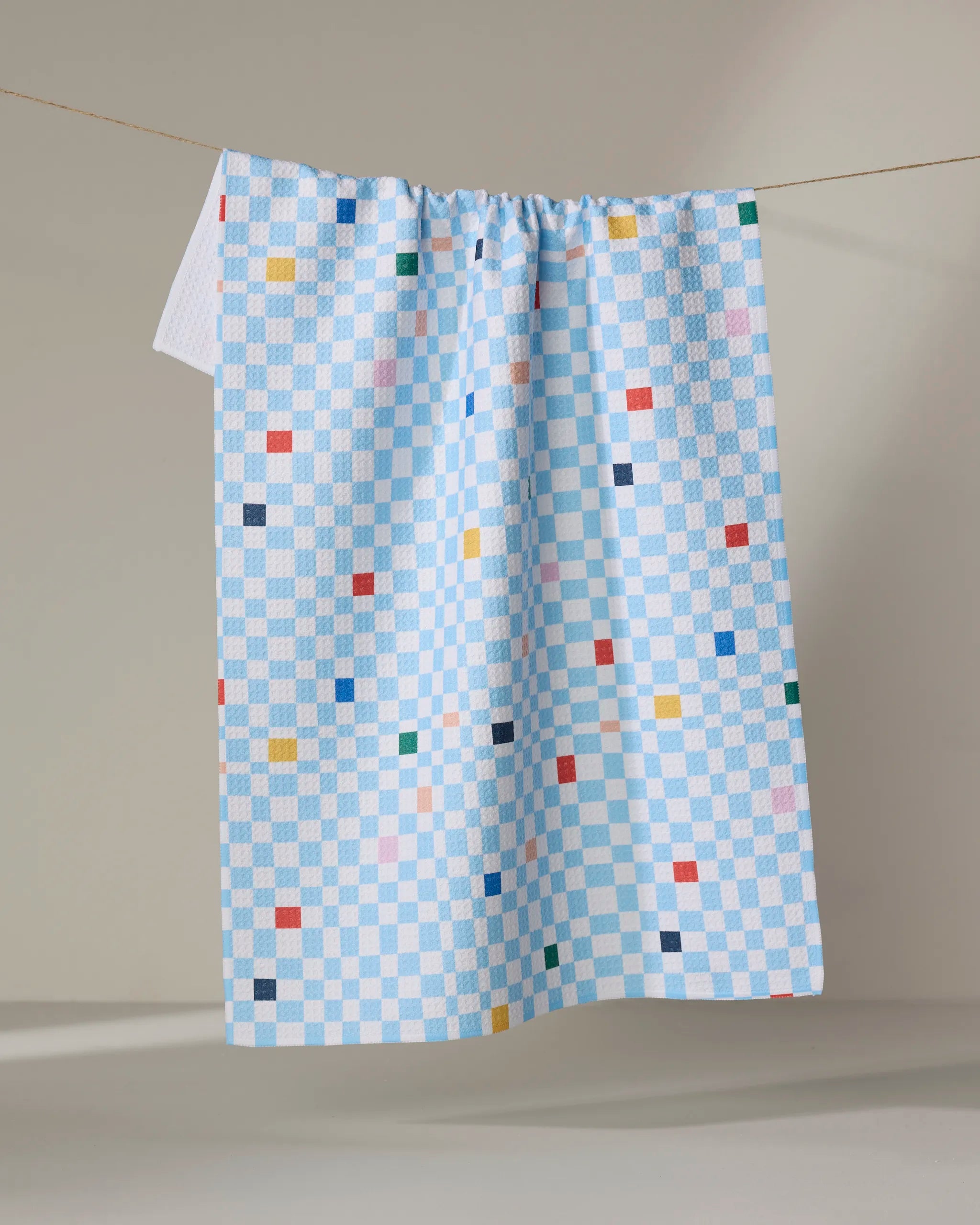 The Happy Hemera "Happy Check" tea towel, featuring blue and white checks with colorful squares and made from recycled materials, hangs on a line against a neutral background.