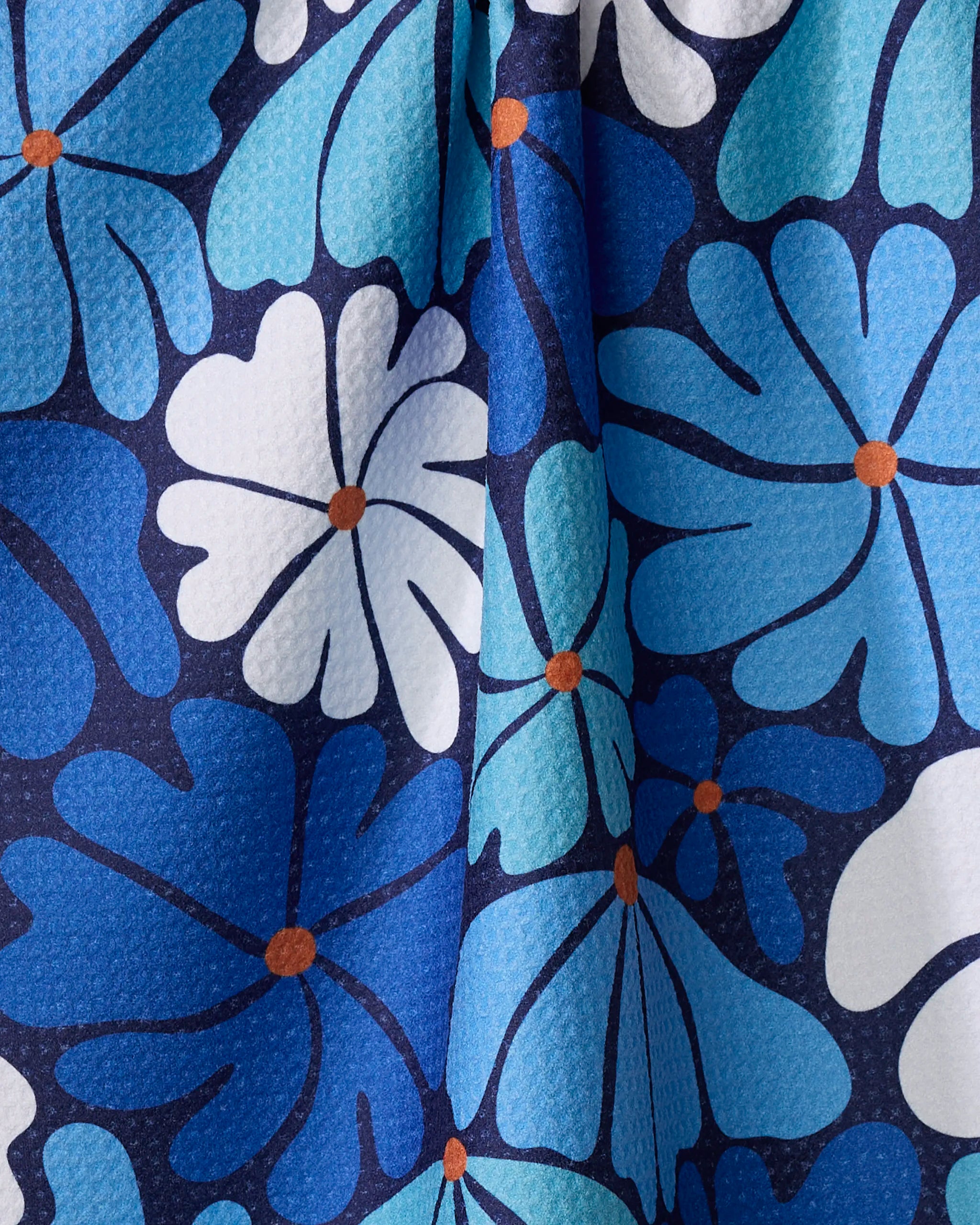 A close-up of the "Happy Fields" recycled kitchen towel by Rachel Parker, showing its textured folds and bold blue, white, and teal floral pattern with orange centers on a dark background.