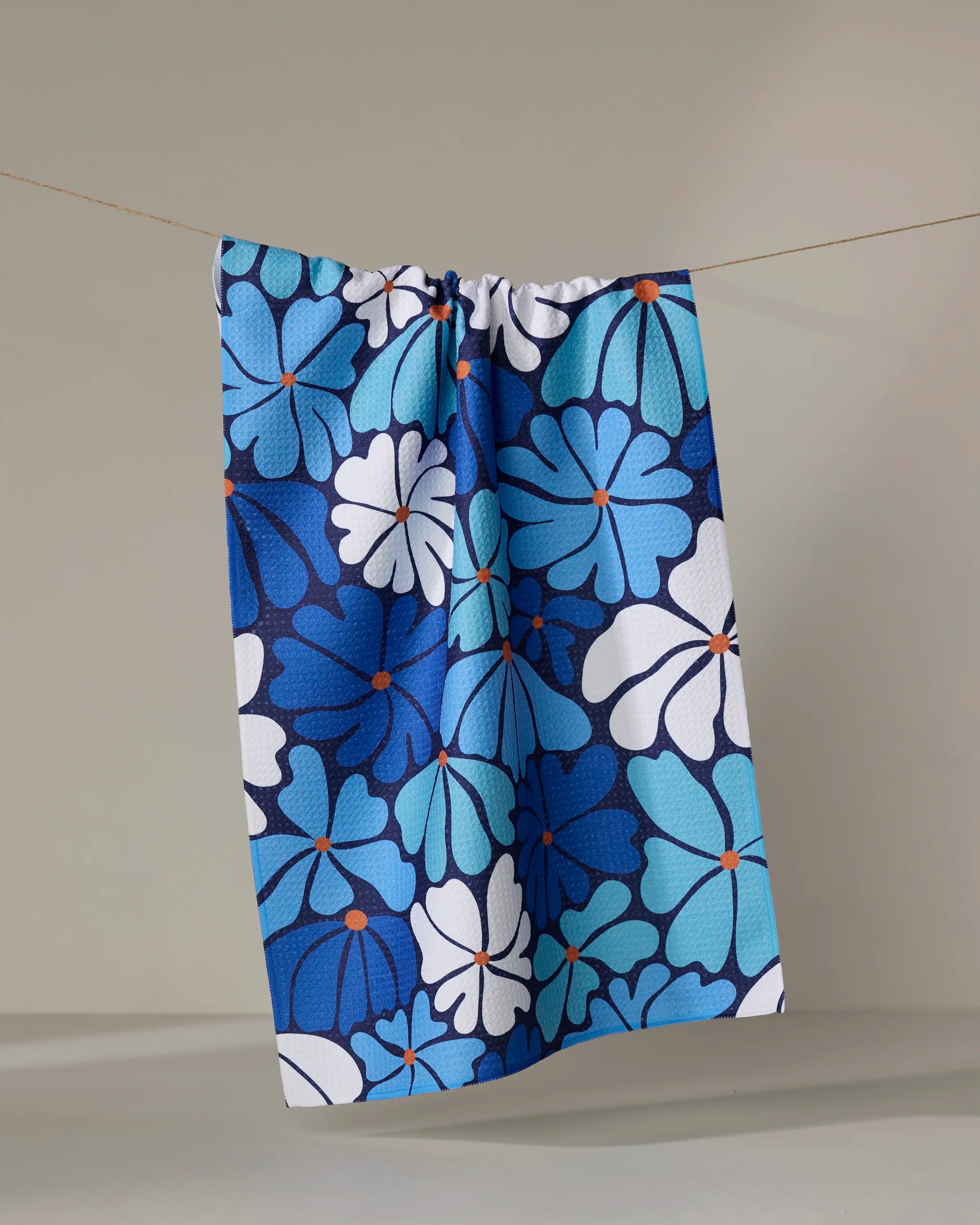 The "Happy Fields" tea towel by Rachel Parker, featuring a blue and white floral pattern with orange accents, hangs from a thin string against a plain light gray background.