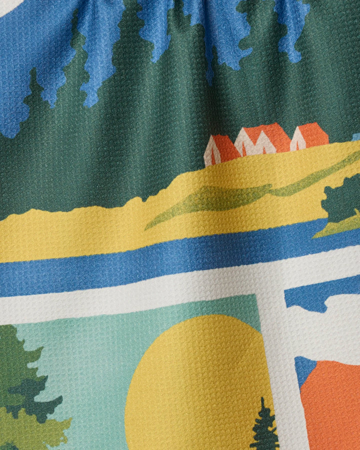 A close-up of Geometry’s “Happy Hiking” eco-friendly tea towel, featuring a vibrant landscape design with trees, houses, hills, and a yellow sun in blue, green, yellow, white, and orange hues.