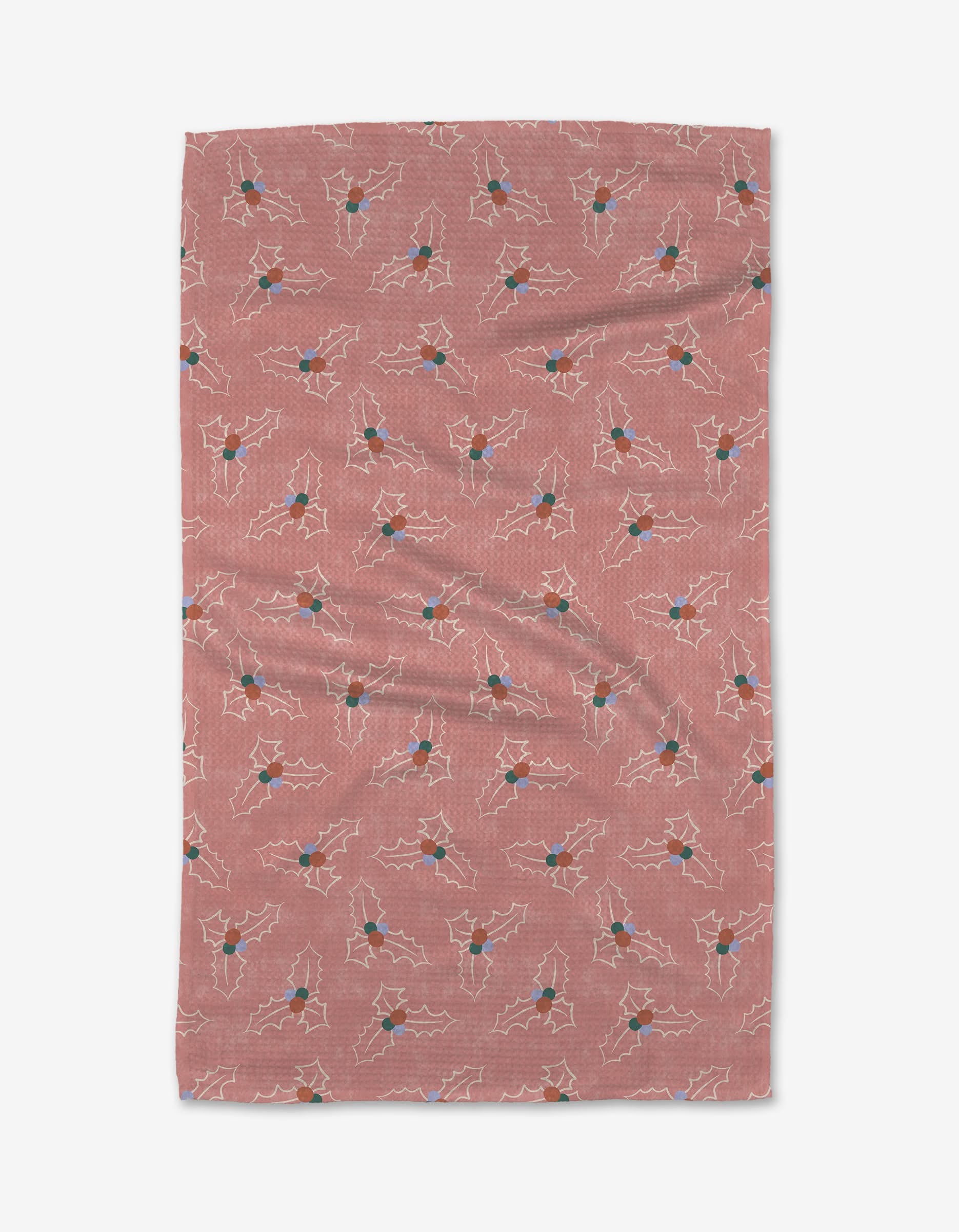 The Happy Holly Berry tea towel by Claire Iglesias features a soft pinkish-red color with green, red, and white holly leaf and berry patterns. This ultra-absorbent towel is displayed flat with slight folds against a white background.