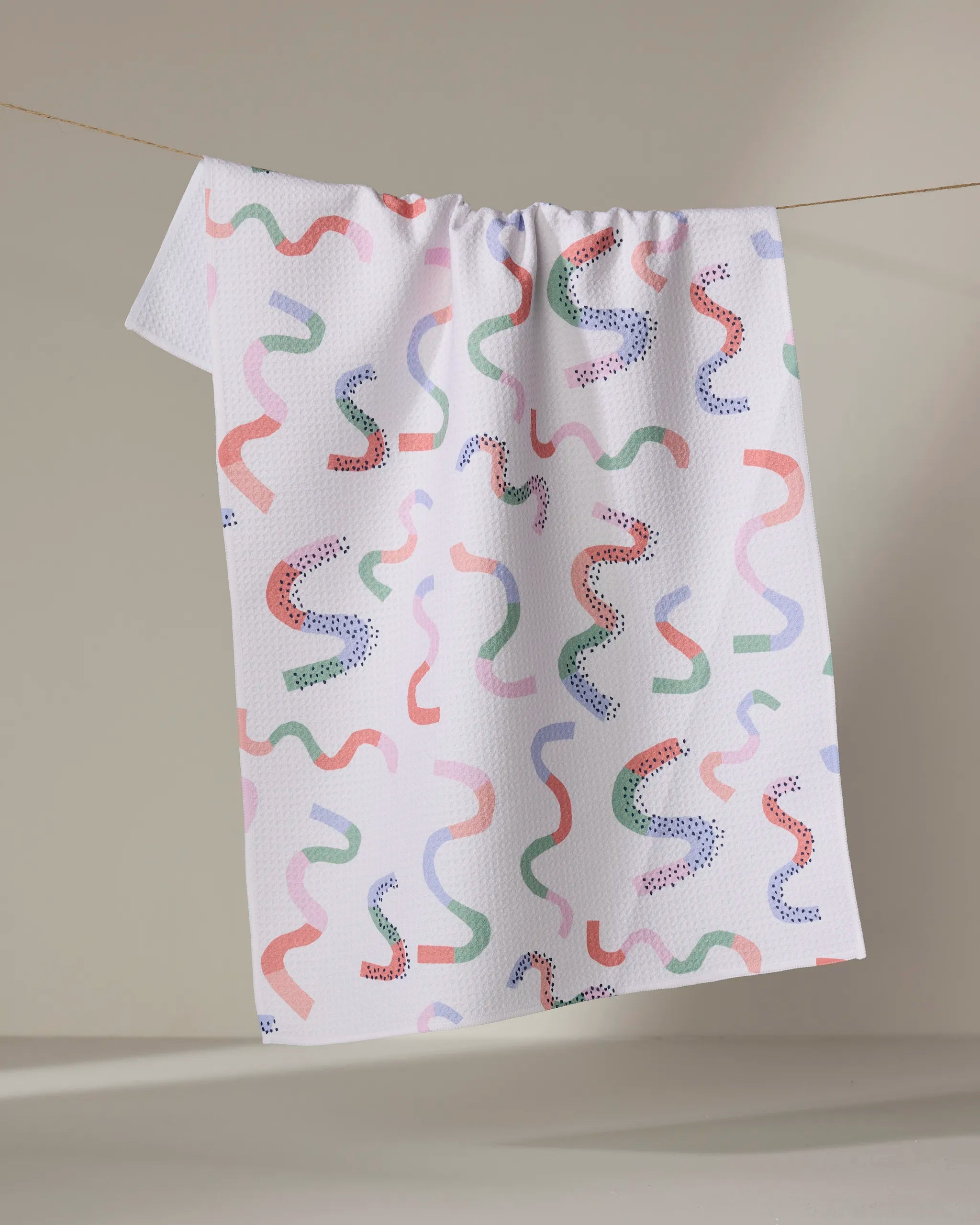 The Claire Iglesias Happy Streamers towel, ultra-absorbent and white with red, green, blue, and purple squiggle and dot patterns, is draped on a clothesline against a neutral background.