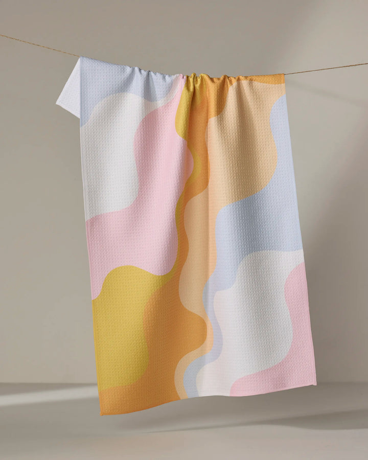 The Gerdadzy Happy Waves dish towel, featuring absorbent pastel fabric with wavy yellow, pink, orange, blue, and white patterns, hangs on a clothesline against a neutral background.