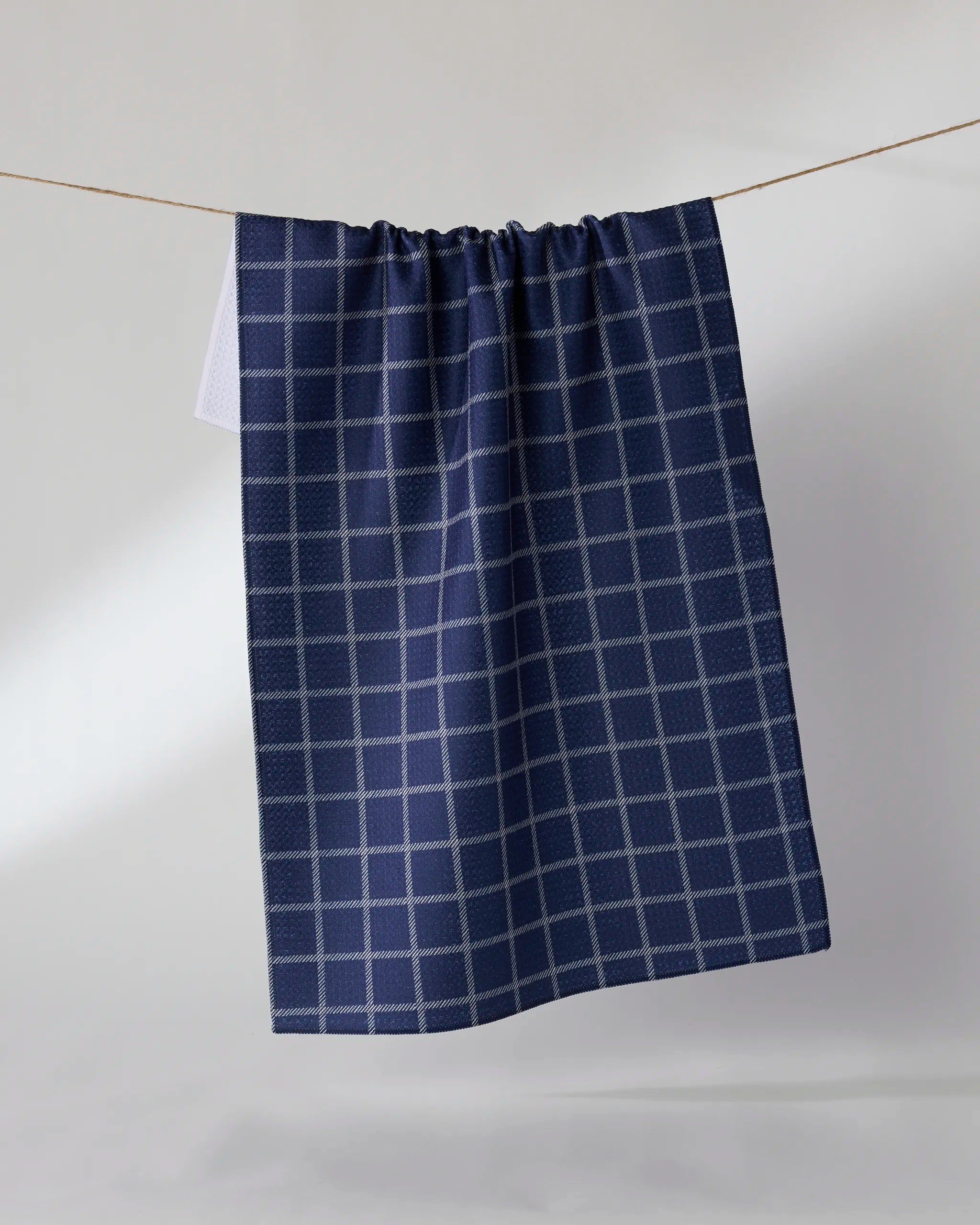 The Geometry Harbor Plaid tea towel, featuring a blue and white checkered design, hangs on a clothesline against a simple light background.