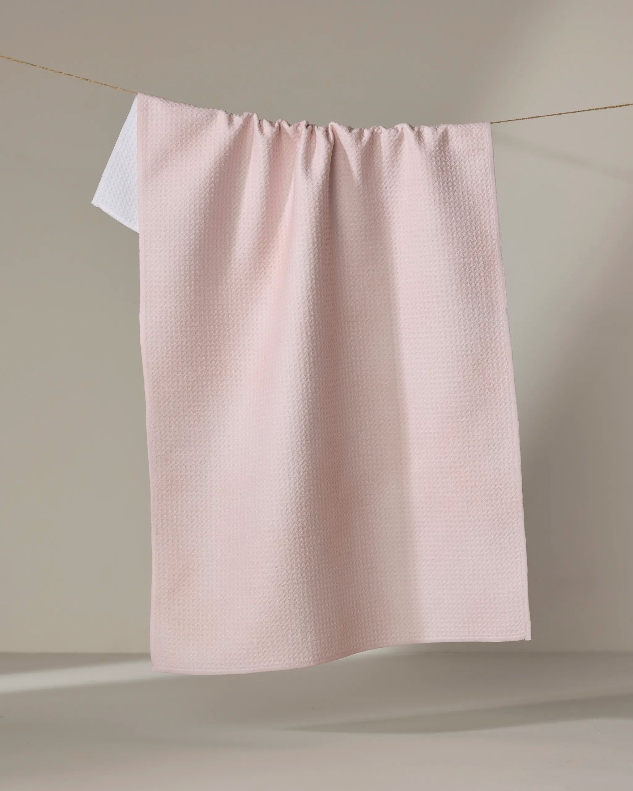 The Harper Pink recycled kitchen towel by Kailee Wright, featuring a light pink textured design, hangs on a clothesline in front of a neutral background with part of a white towel peeking from behind.