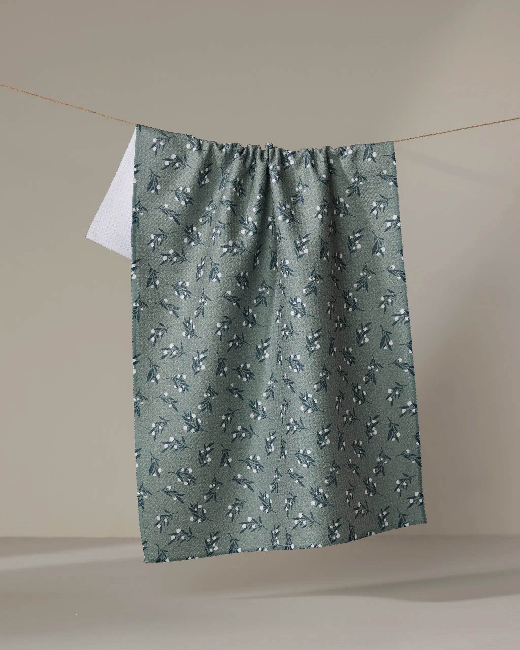 The "Harvest Berries" recycled kitchen towel by Charlie Rowan Designs, featuring a green leaf and berry pattern, hangs on a clothesline with its white reverse side showing against a plain beige background.