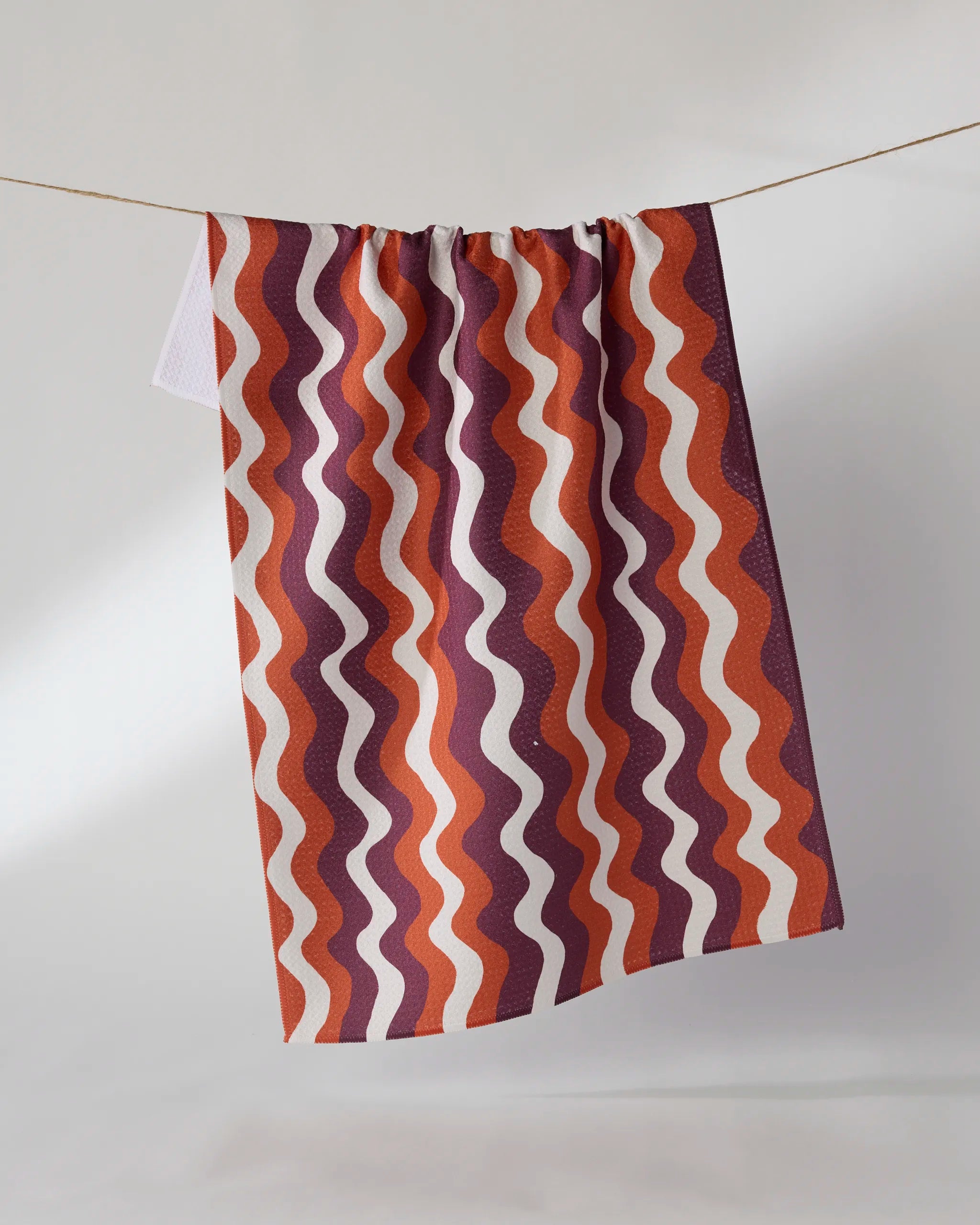 The Geometry Harvest Groove kitchen tea towel, featuring wavy maroon, orange, and white stripes, hangs from a clothesline against a plain light background.