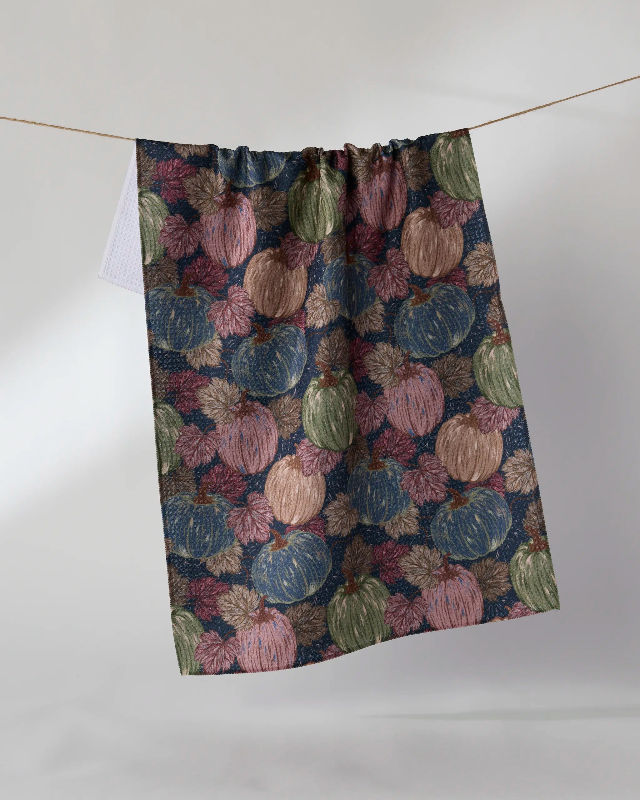 The Geometry Harvest Moon kitchen towel, made from eco-friendly materials, hangs on a line and displays pumpkins and leaves in muted blue, green, pink, and brown tones set against a dark background.