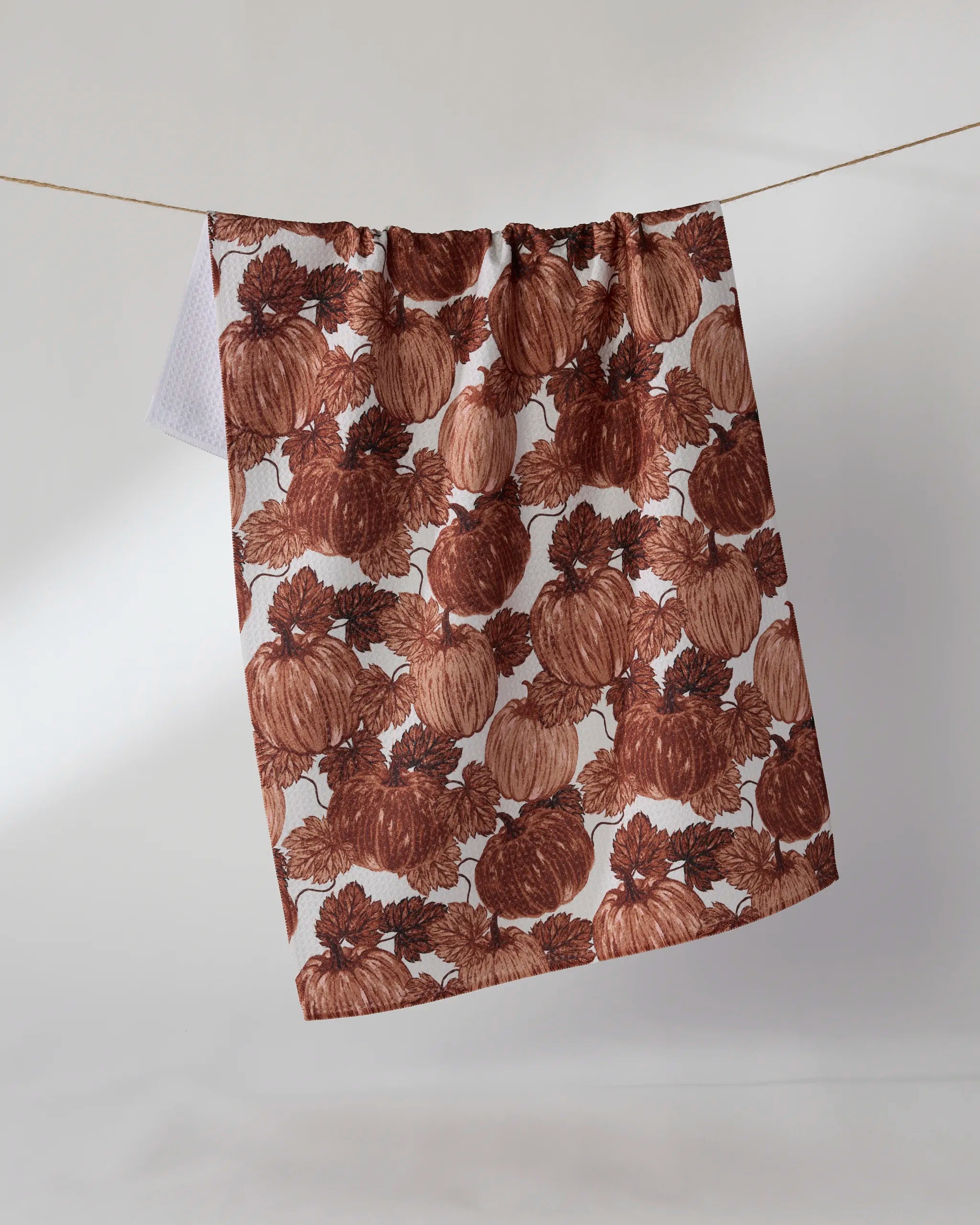 The Geometry Harvest Moon eco-friendly kitchen towel, featuring a brown pumpkin and leaf pattern, hangs on a clothesline against a plain, light background.