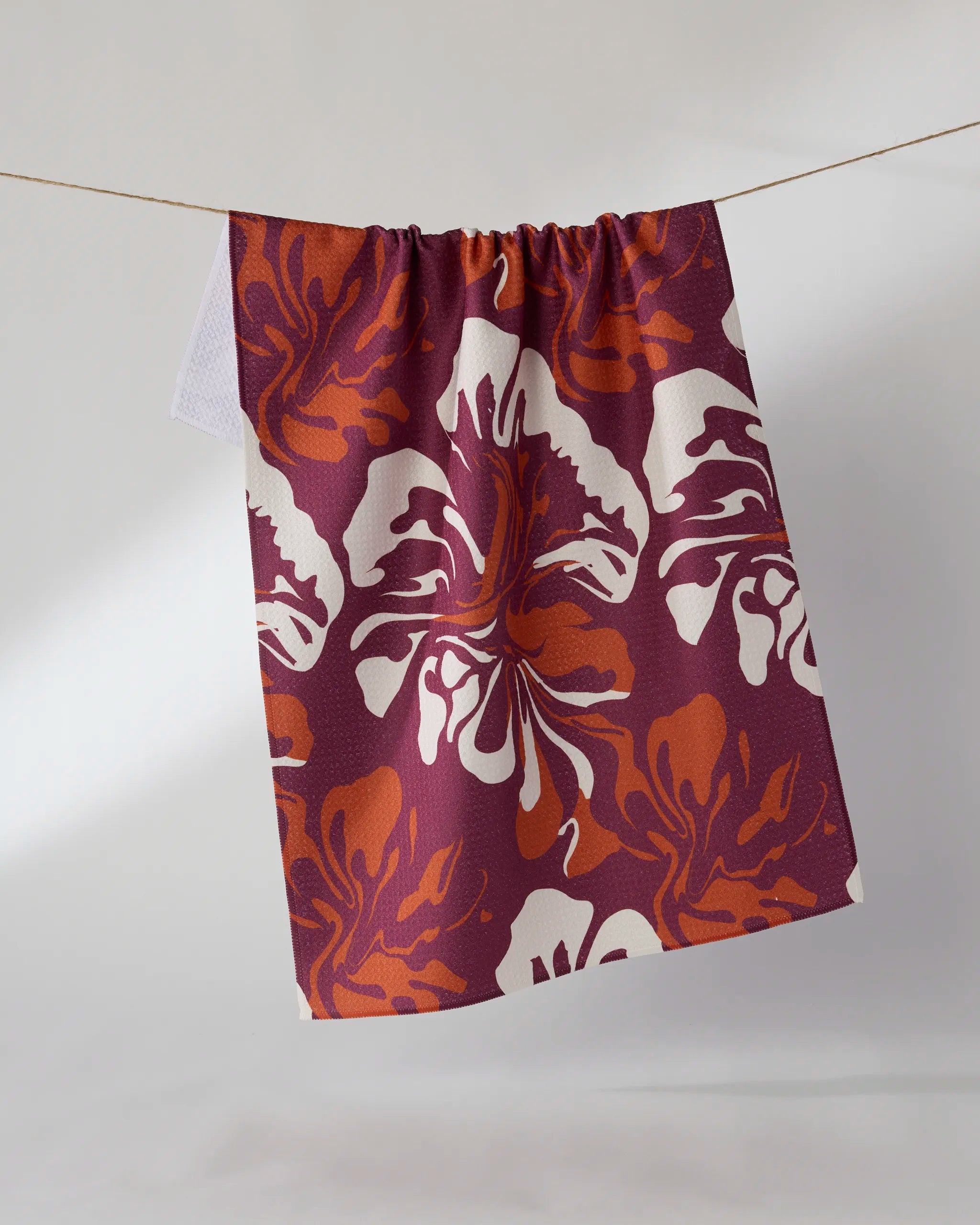 The Geometry Harvest Muse tea towel, ultra-absorbent and featuring a bold white and orange floral pattern on deep maroon, hangs on a clothesline against a plain light background.