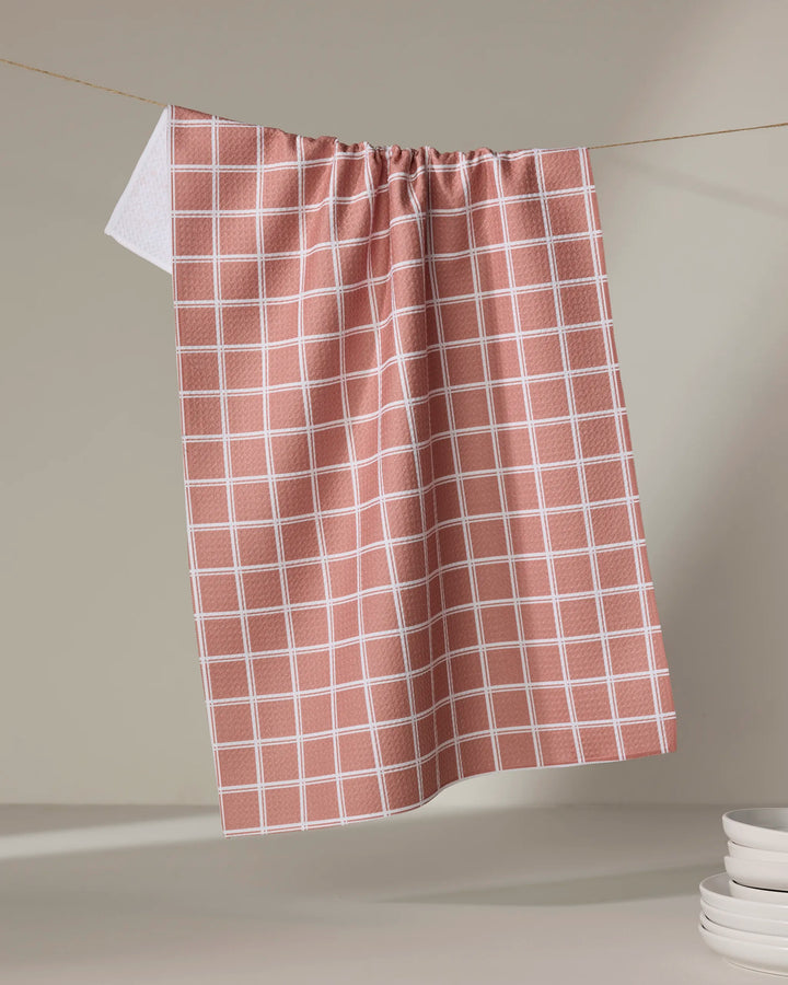 The Harvest Plaid tea towel by Charlie Rowan Designs hangs on a string against a neutral background, with a stack of white plates partially visible in the lower right corner.