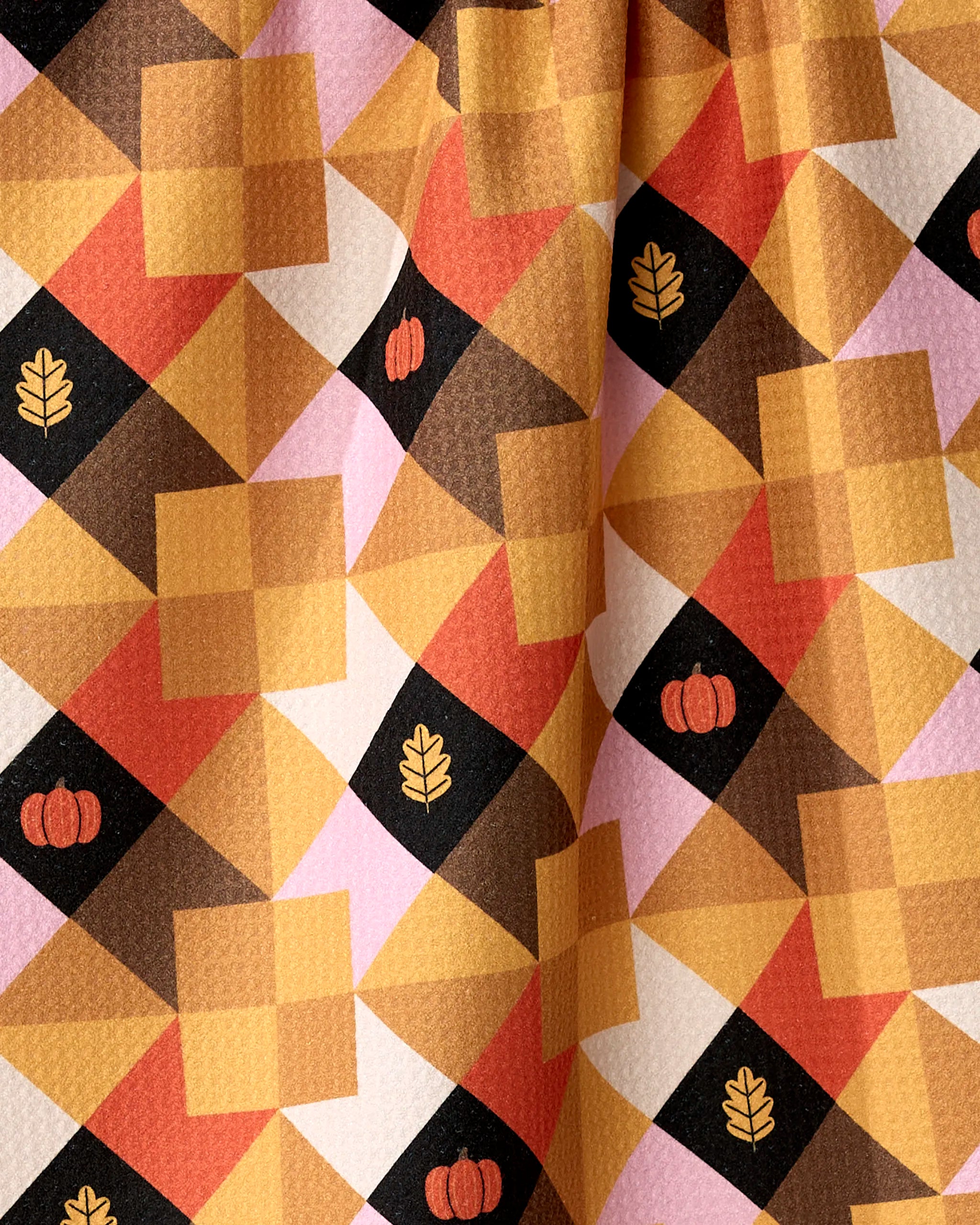 The Harvest Quilt tea towel by Megan Roy is made from recycled materials and showcases a festive autumn geometric pattern in orange, brown, yellow, and pink with diamonds, squares, pumpkins, and leaf icons.