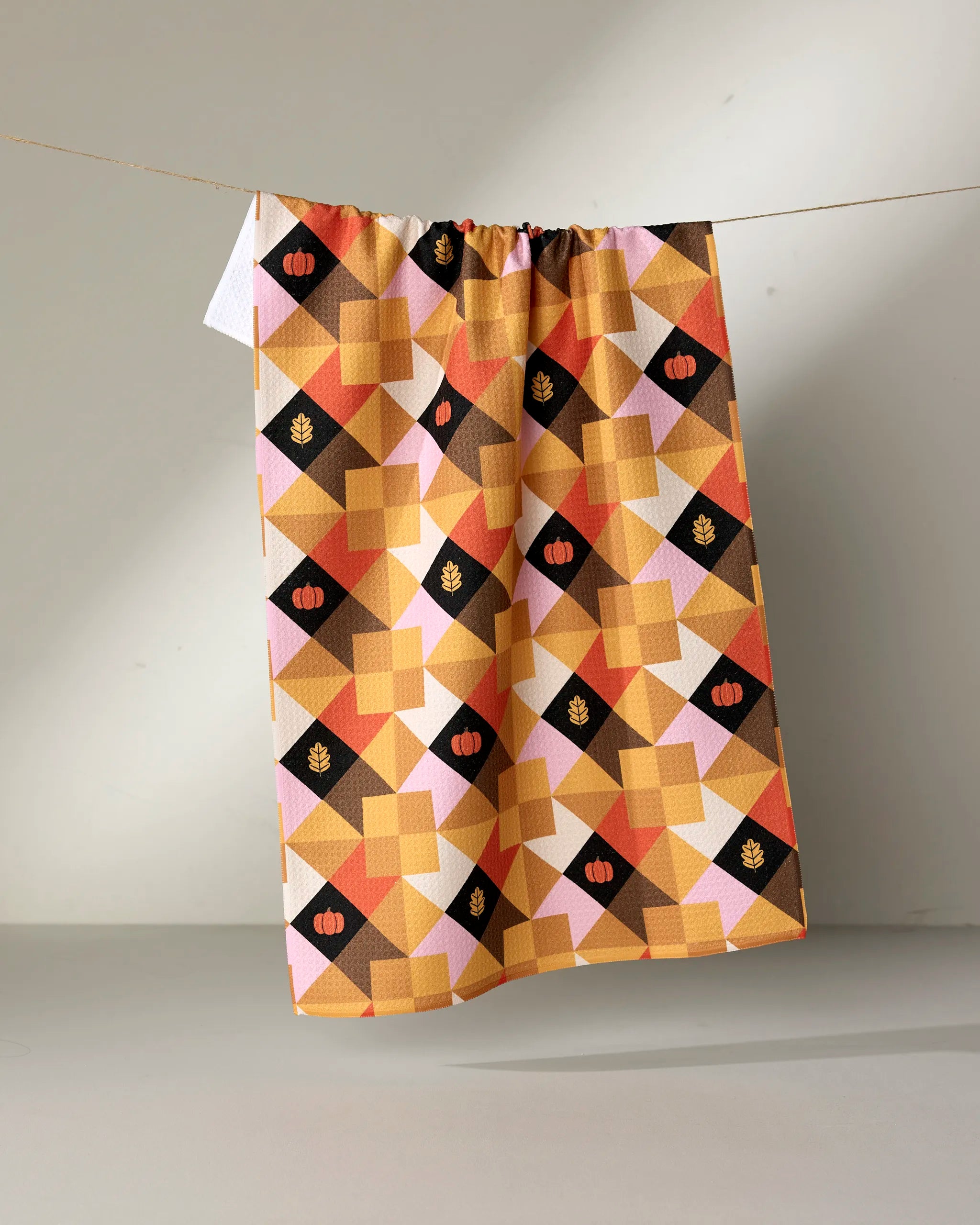 The Megan Roy Harvest Quilt kitchen tea towel, made from recycled materials and featuring an orange, brown, and yellow geometric patchwork with pumpkins and leaves, hangs on a clothesline against a neutral backdrop.